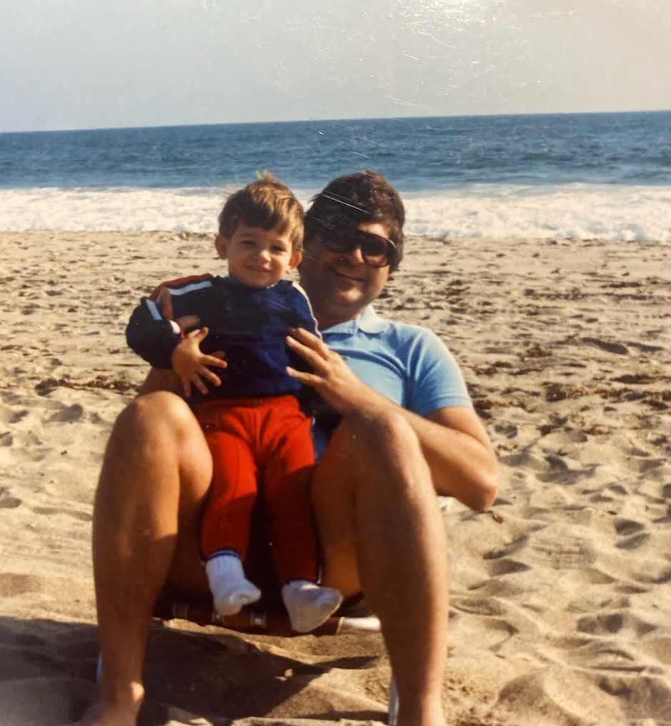 A man and a young boy sitting on a beach with the ocean in the background. The man is wearing sunglasses and a light blue shirt, and the boy is wearing a navy blue jacket and red pants.