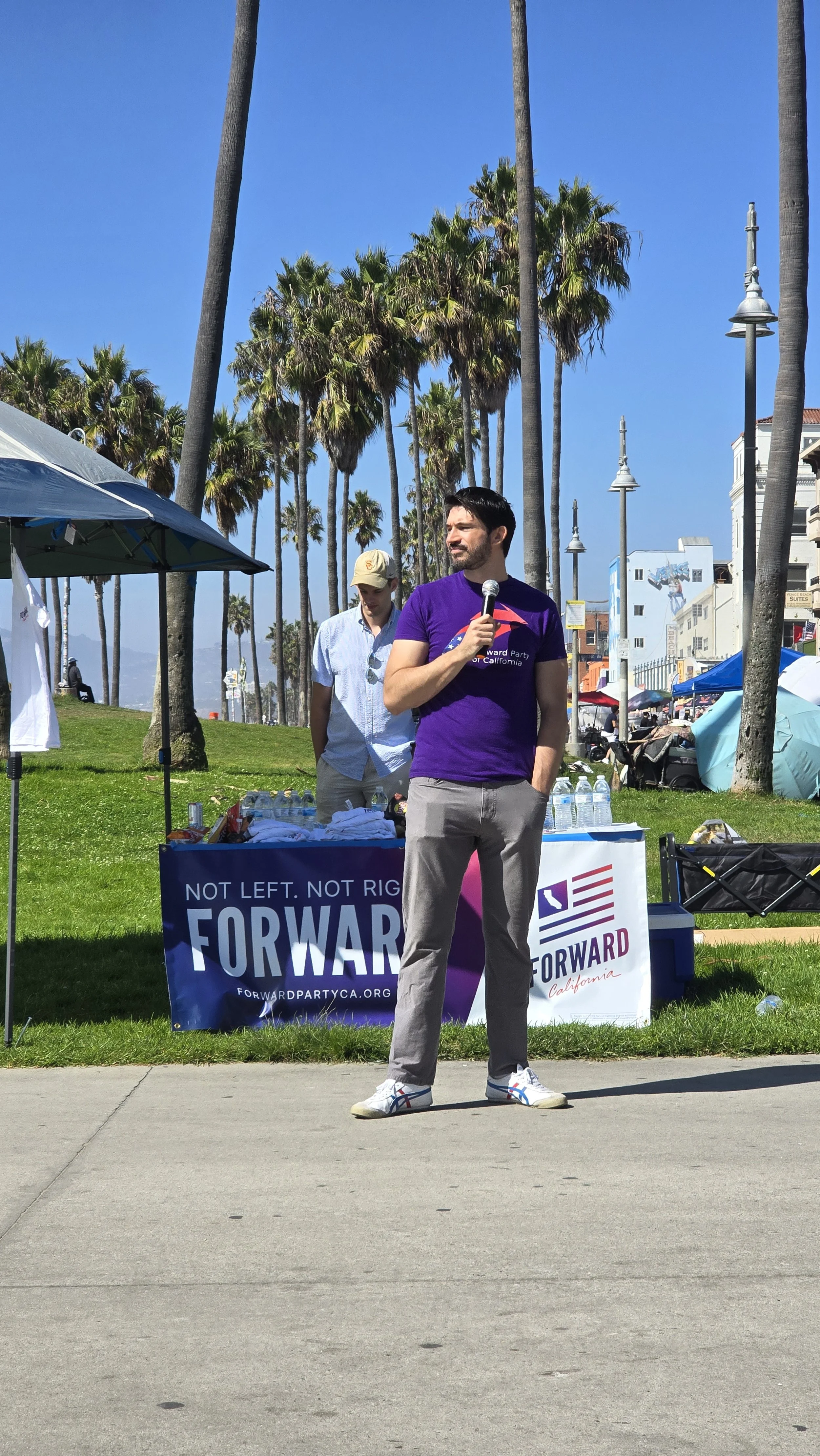 A man holding a microphone standing outdoors in front of a booth at a political event, with tall palm trees and a clear blue sky in the background.