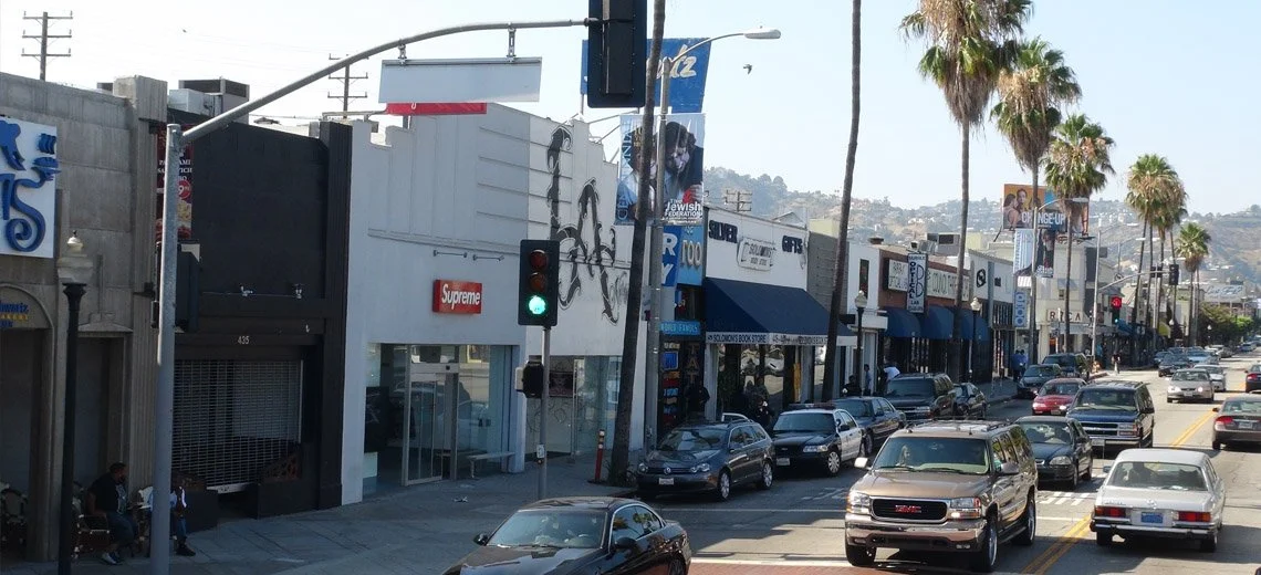 Street scene with parked cars, palm trees, and storefronts including a Supreme store, under a bridge, with hills in the background.