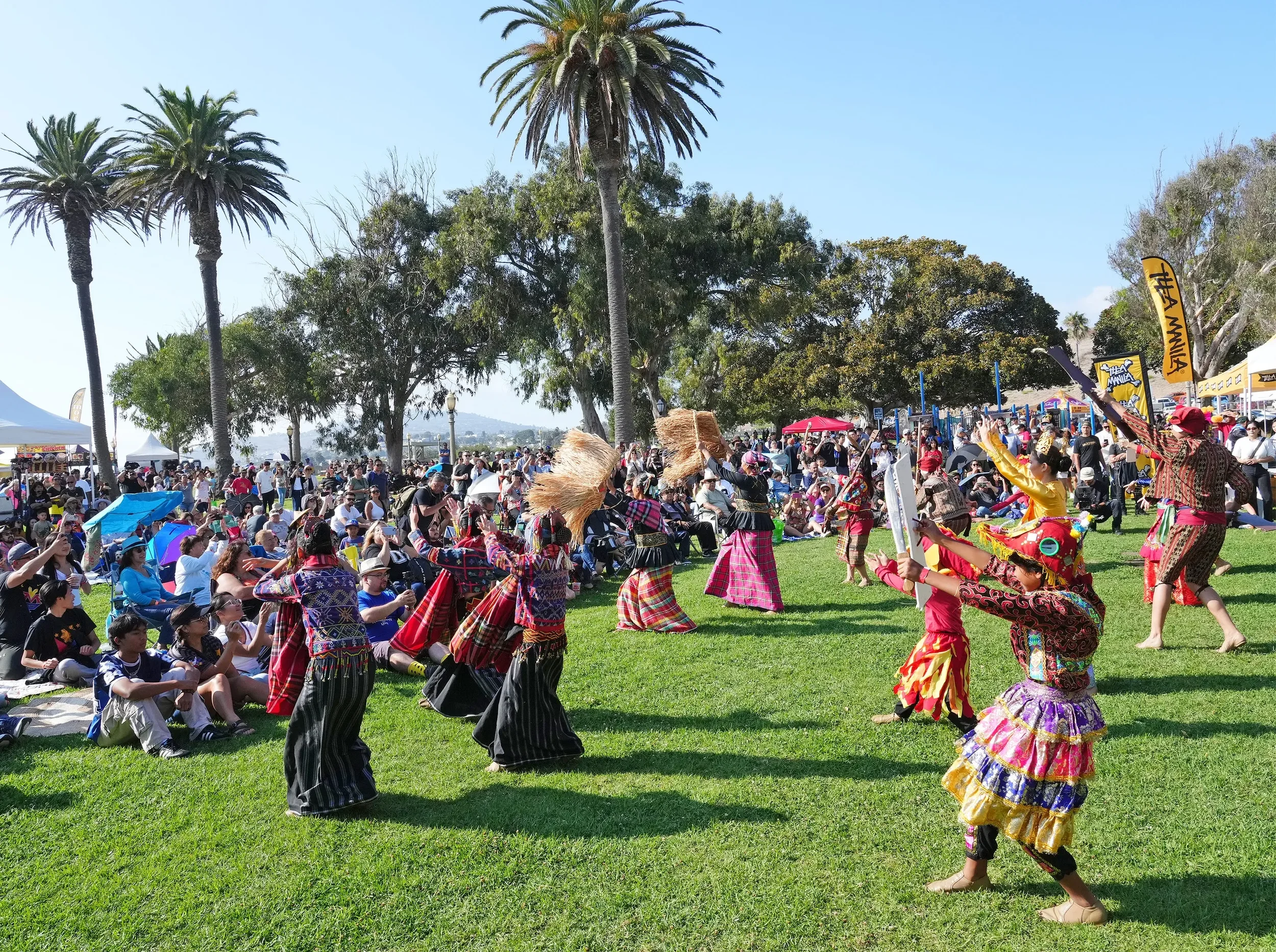 Colorful traditional dancers perform at an outdoor cultural festival in front of a large audience seated on the grass, with palm trees and festival booths in the background.