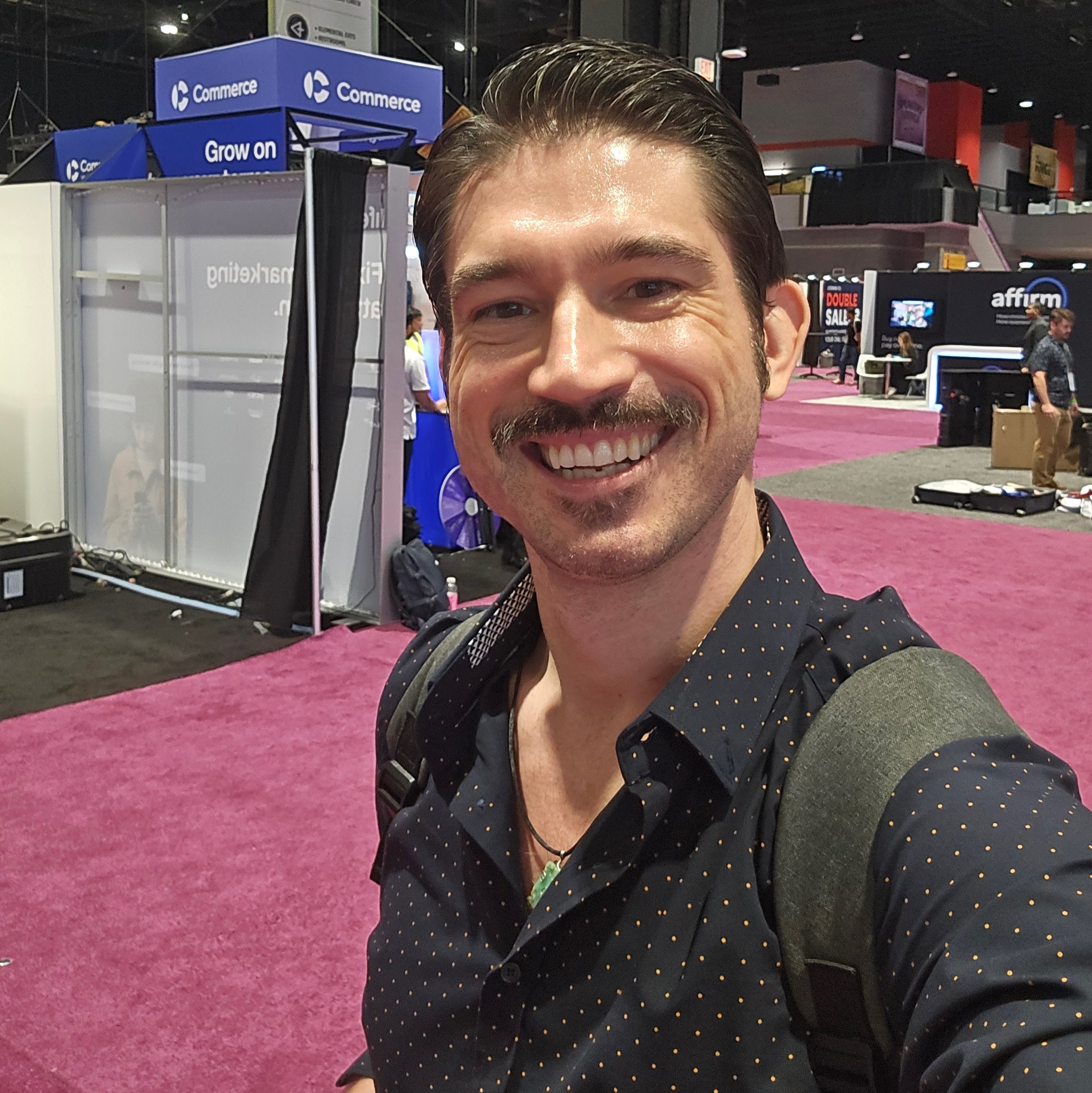 A smiling man taking a selfie at a trade show or convention, with booths and displays in the background, including signs for 'Commerce' and 'Affirm', on a pink carpeted floor.