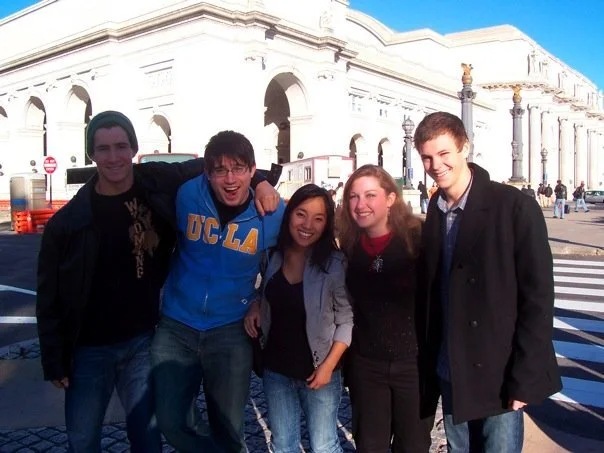 Group of five young friends smiling and standing together outdoors in front of a large white building with arches, under a clear blue sky.