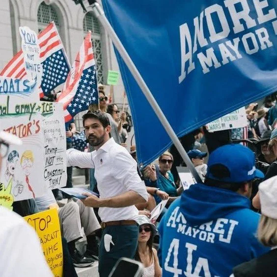 Protesters gather holding signs and flags, including blue 'A MORE MAYOR' flags and American flags, during a rally or demonstration.