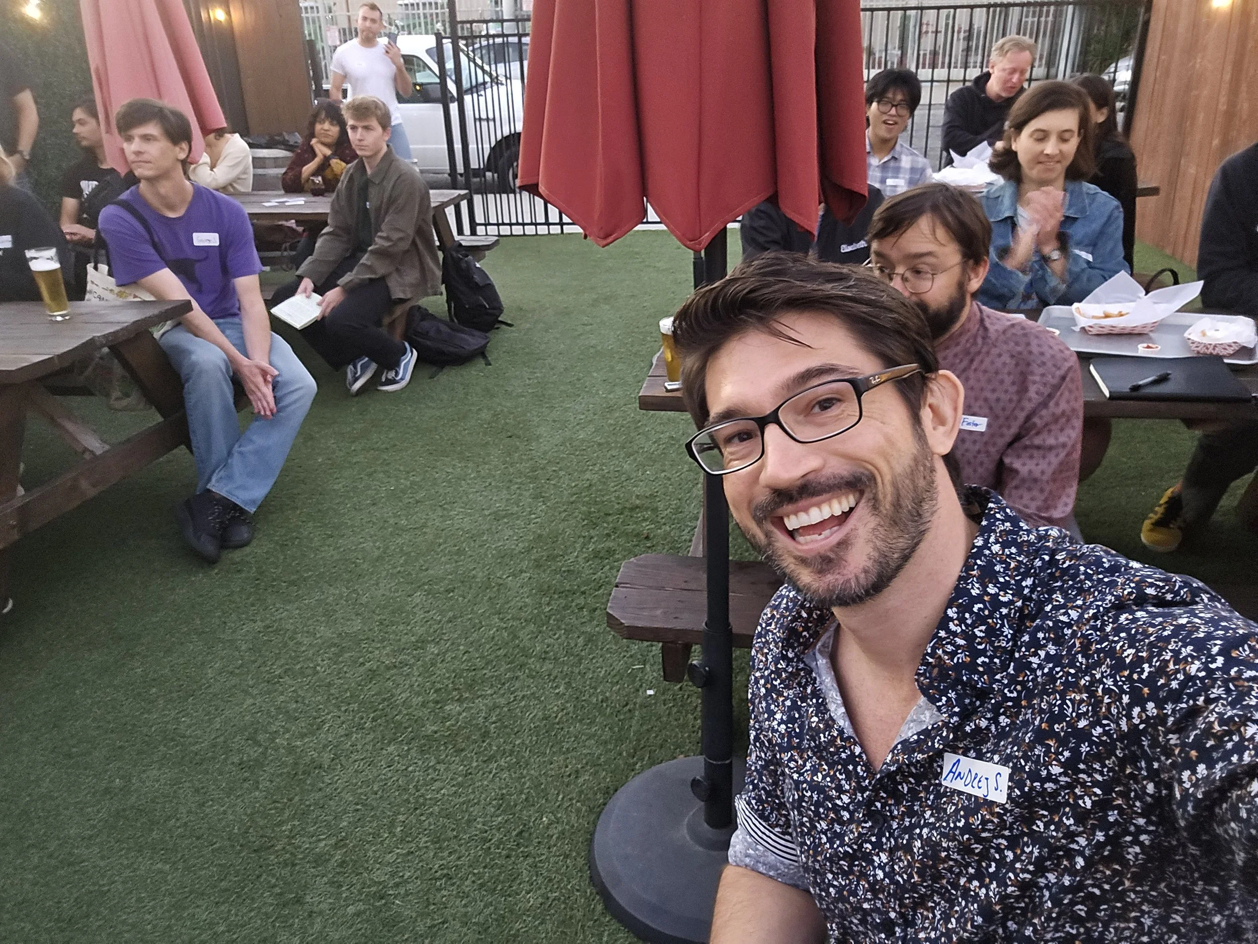A group of people at an outdoor gathering, some sitting at picnic tables and others standing, with a smiling man in the foreground taking a selfie.