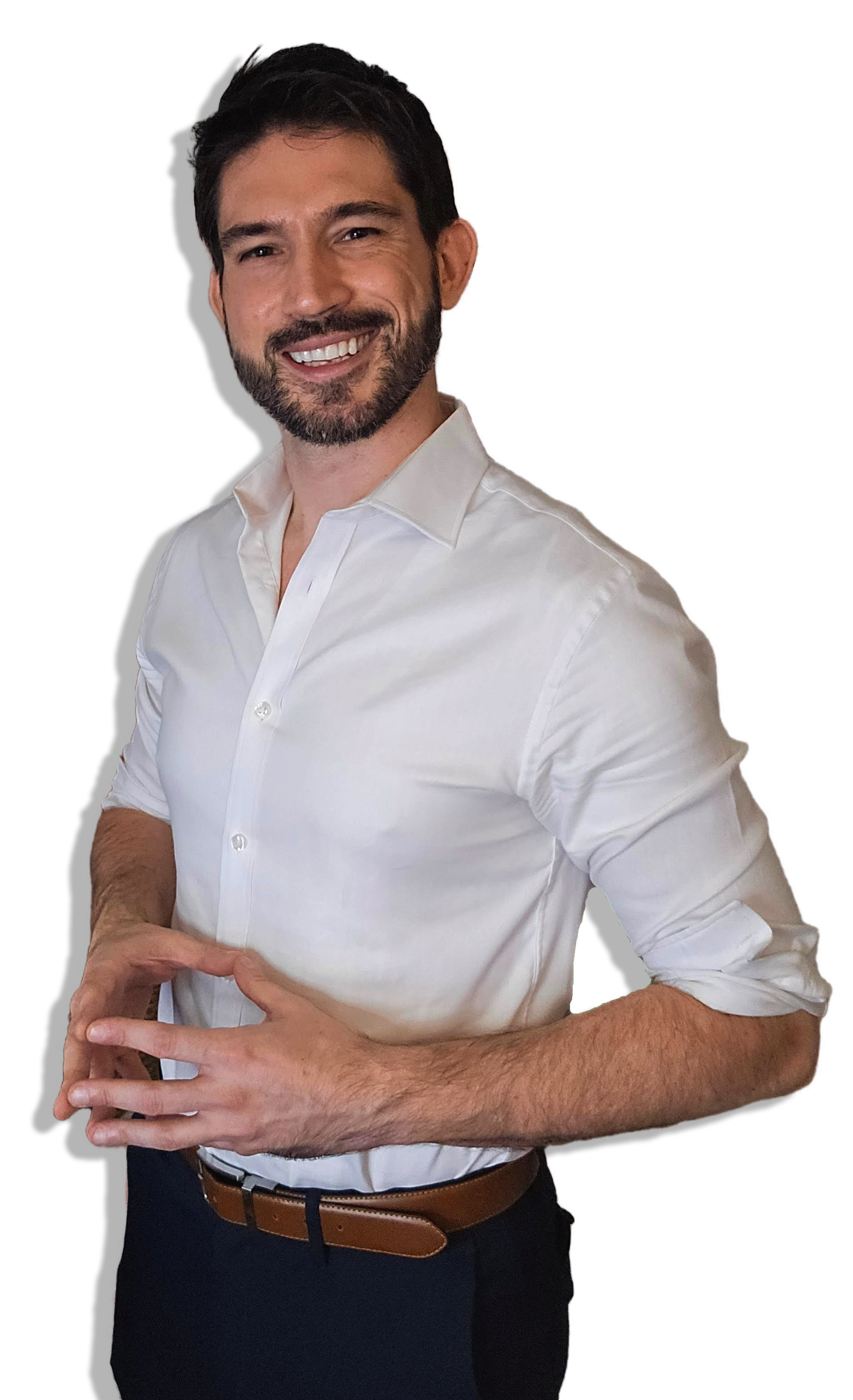A smiling man with dark hair, a beard, wearing a white dress shirt with sleeves rolled up, and dark pants veiled by a brown belt, standing with his hands clasped in front of him.