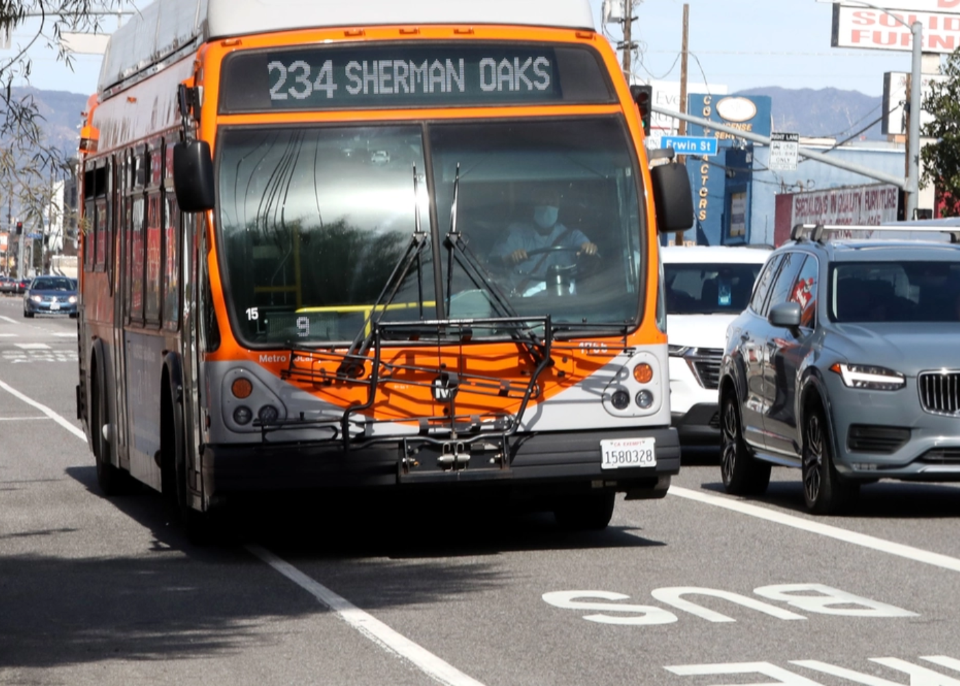 Orange and gray bus with route number 234 to Sherman Oaks on a city street, with cars parked and driving nearby, and buildings in the background.
