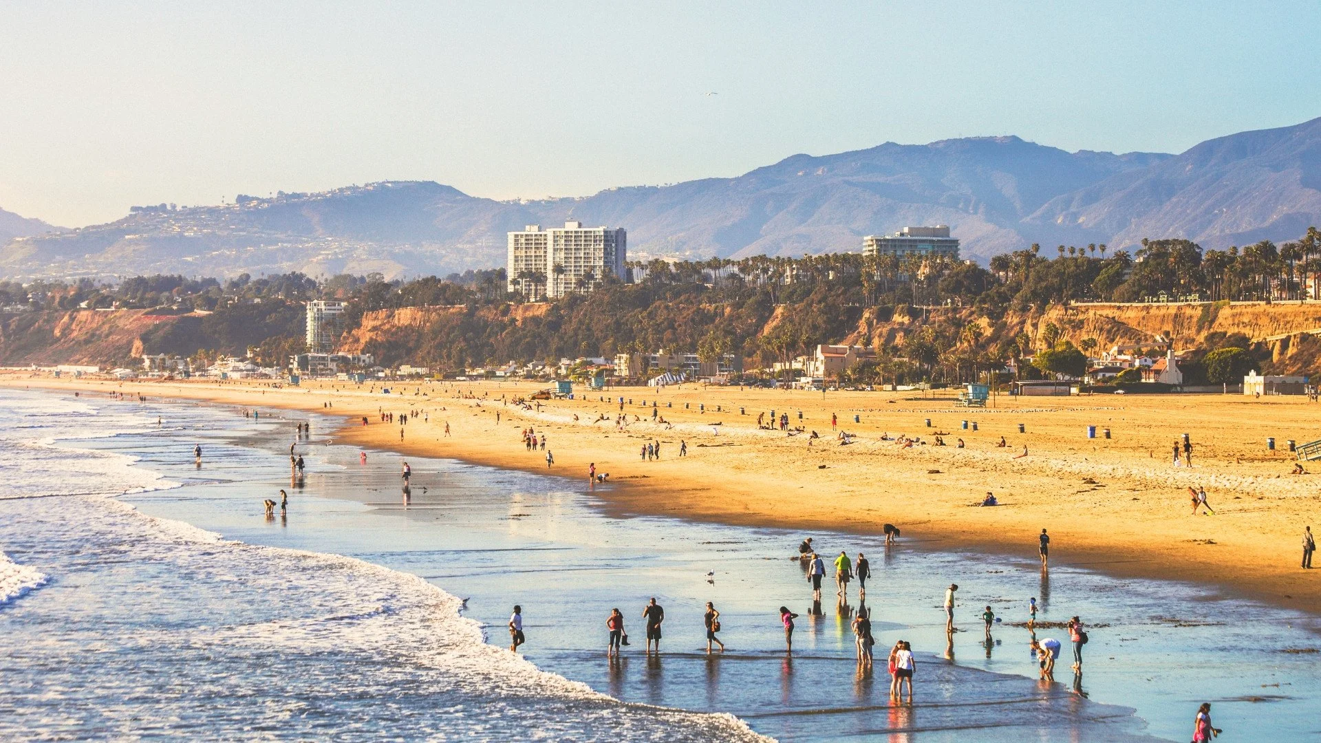 People walking and relaxing on a sandy beach with waves, buildings, and mountains in the background during daytime.