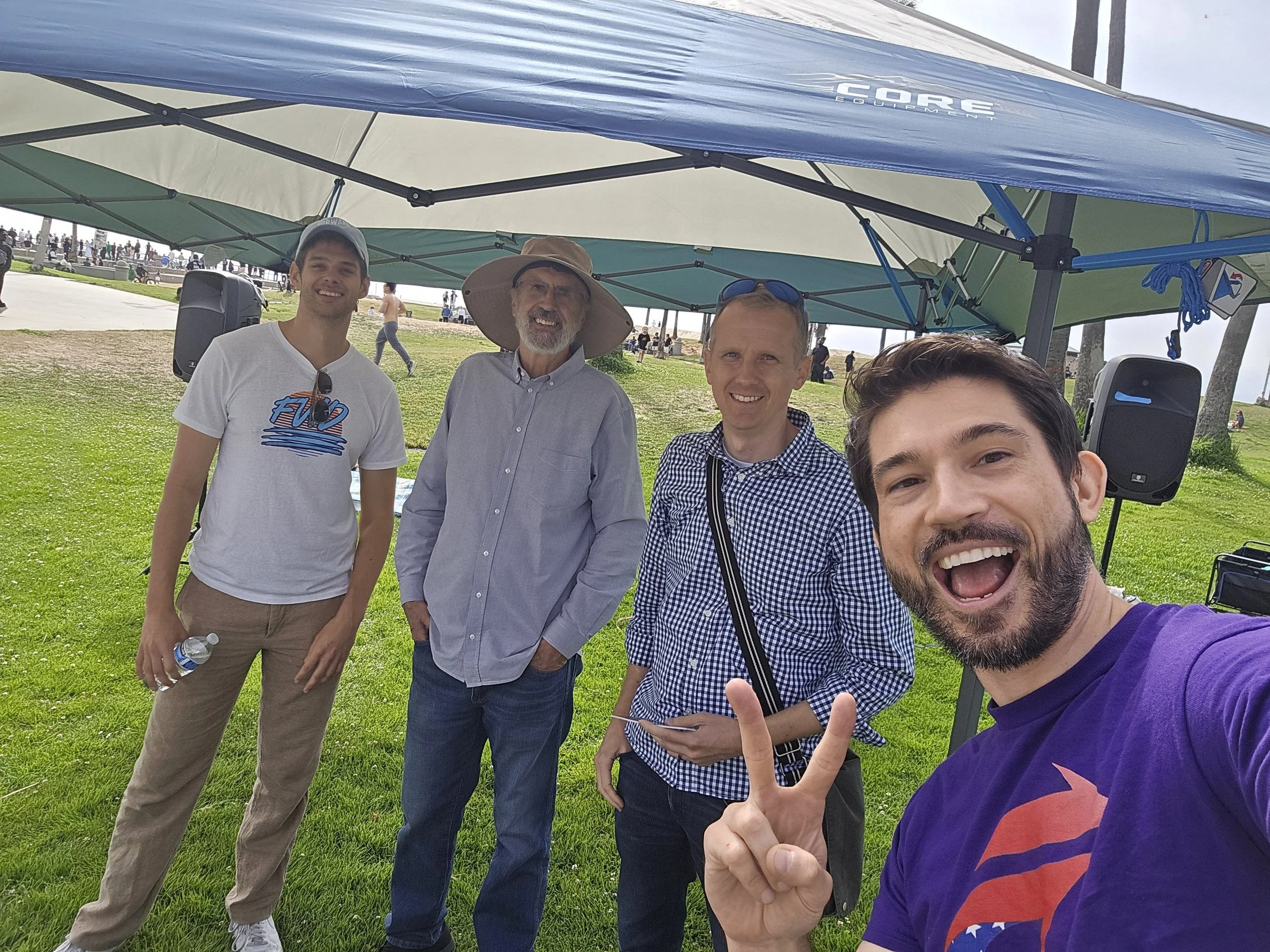 Four men smiling and posing for a photo under a canopy at an outdoor event. The man in the front is making a peace sign.