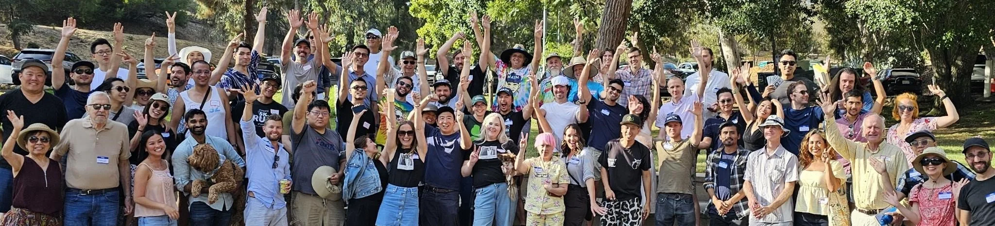 A large group of people outdoors, smiling and waving at the camera, with trees and parked cars in the background.