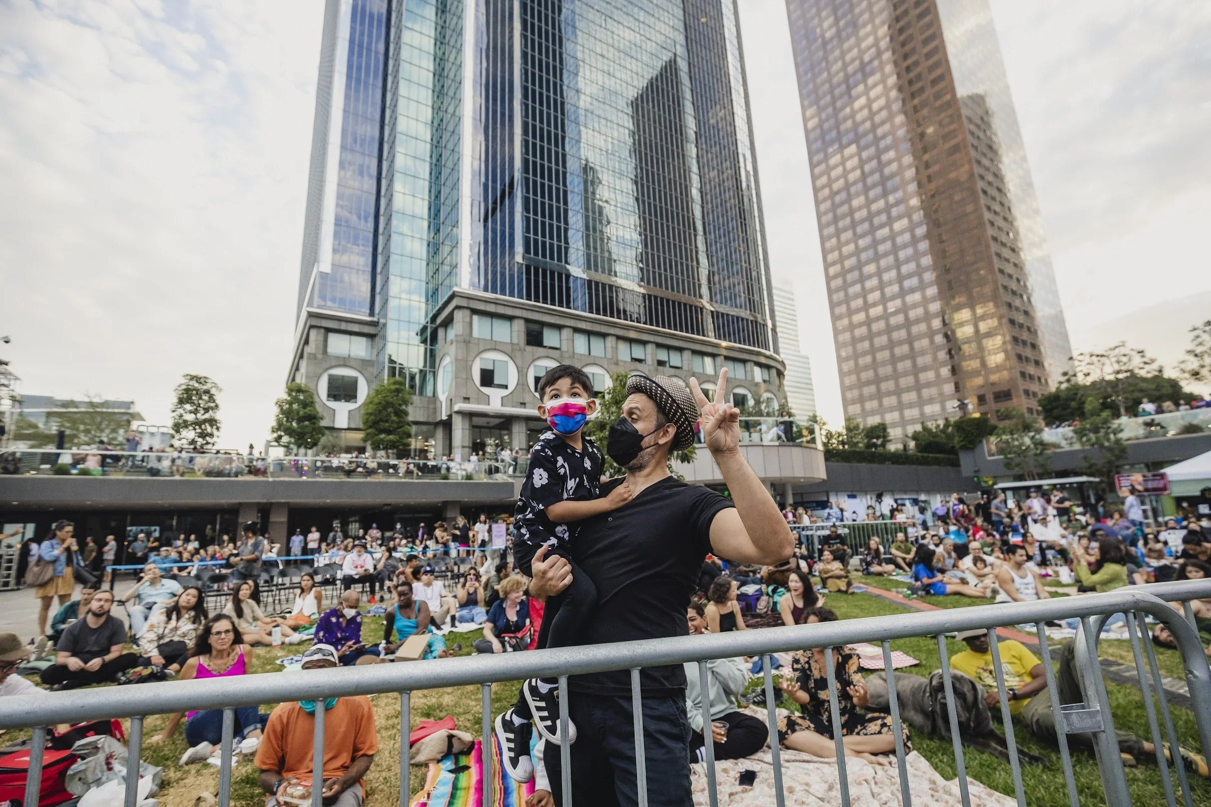 A woman holding a young boy wearing a colorful face mask at an outdoor event in a city. They are surrounded by people sitting on the grass, with tall office buildings in the background. The woman is making a peace sign with her hand.