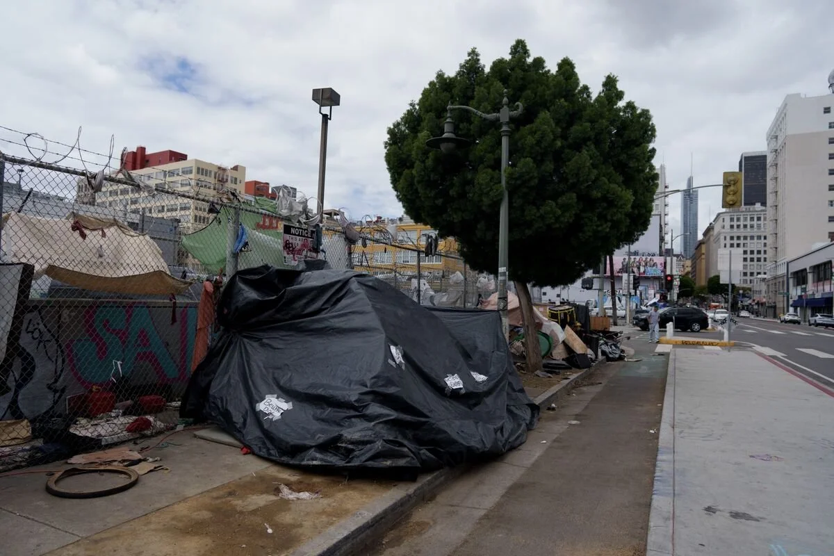 Urban street scene with a fenced area, a large tree, and a covered pile of belongings on the sidewalk.