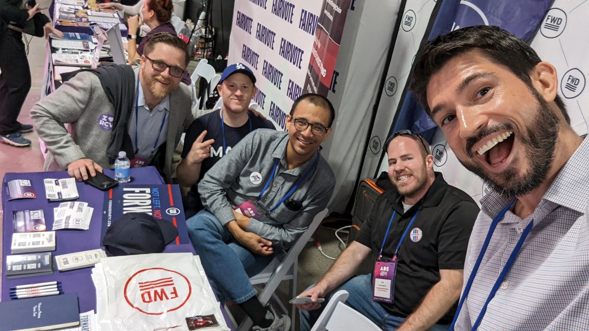 A group of five men at a conference, sitting and standing around a booth with promotional materials. One man is taking a selfie, all smiling.