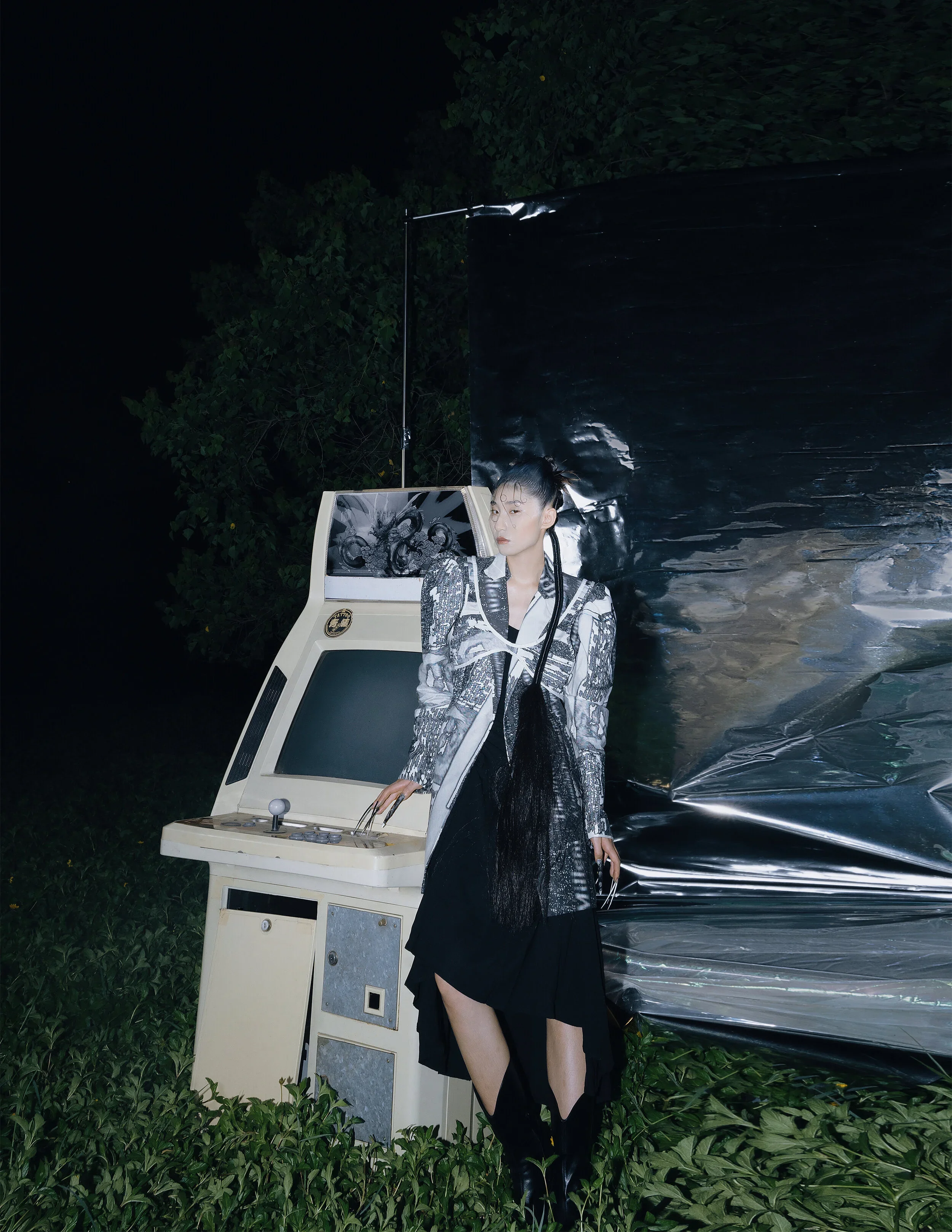 A woman standing outdoors at night post-fashion photoshoot, next to an arcade machine, with metallic foil and dark greenery in the background.