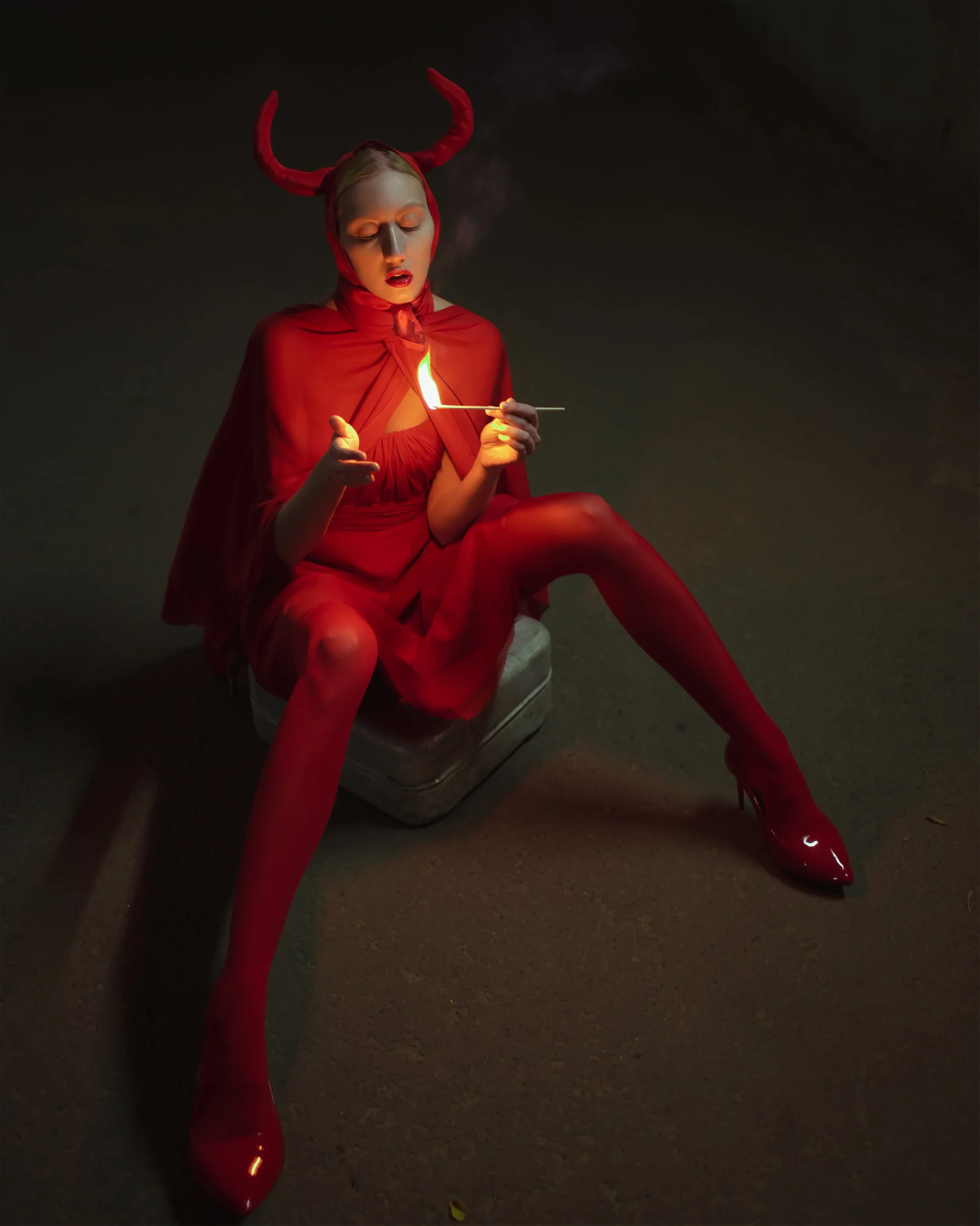 Person dressed as a devil sitting on a small stool, wearing a red outfit with a cape, horns, and red high heels, lighting a match with a flame.