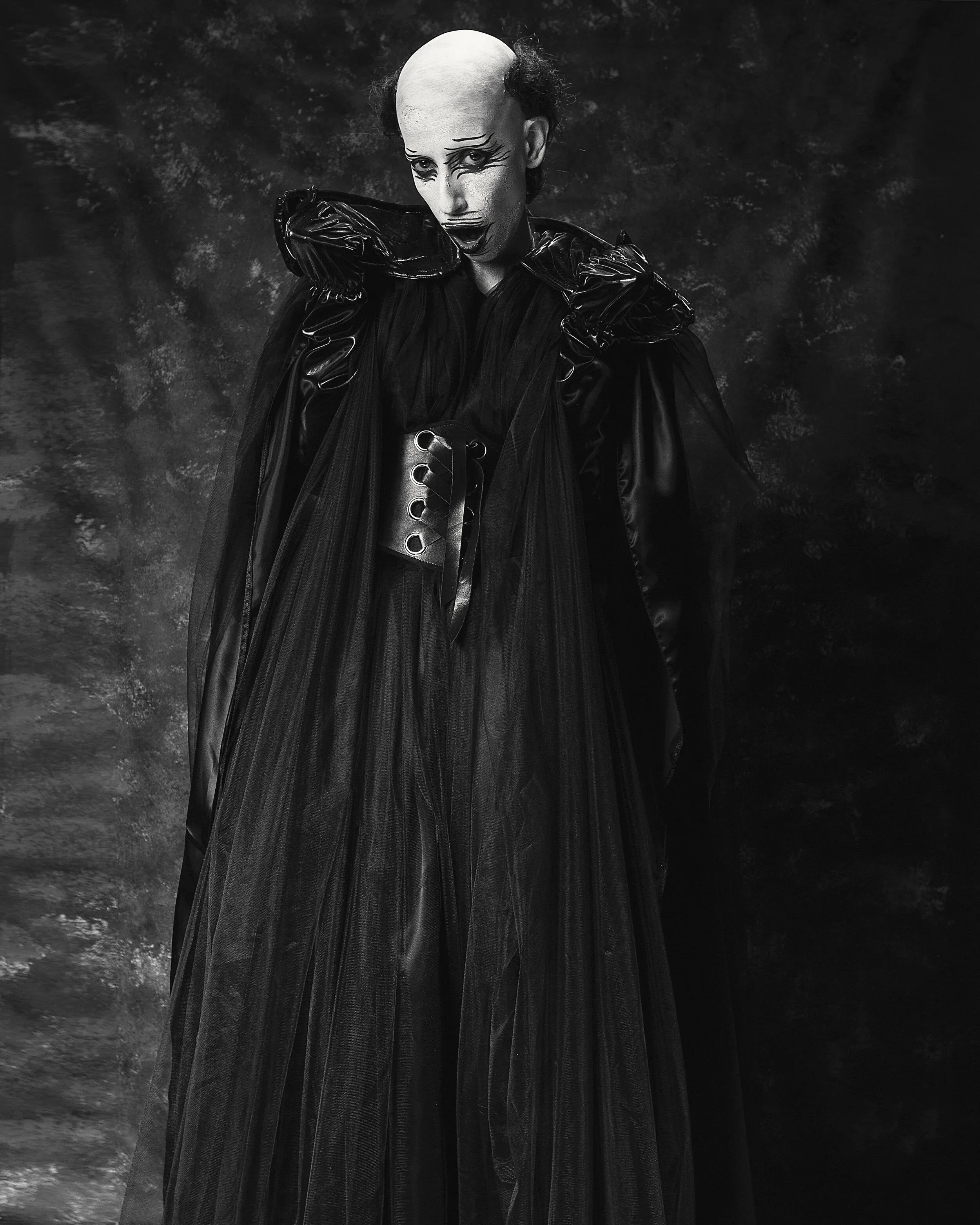 A person dressed in a black, gothic-style costume with dramatic makeup and a bald cap, posing against a dark, textured background.