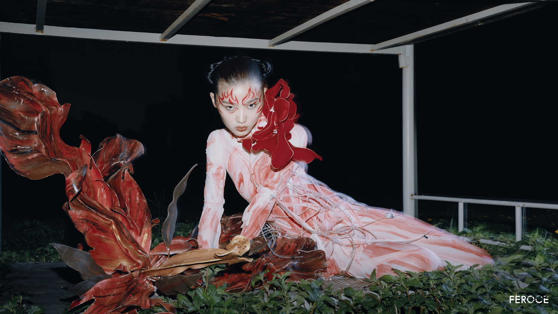 A person with elaborate makeup and a flowing pink and red costume poses on the ground under a structure at night, surrounded by large decorative leaves.