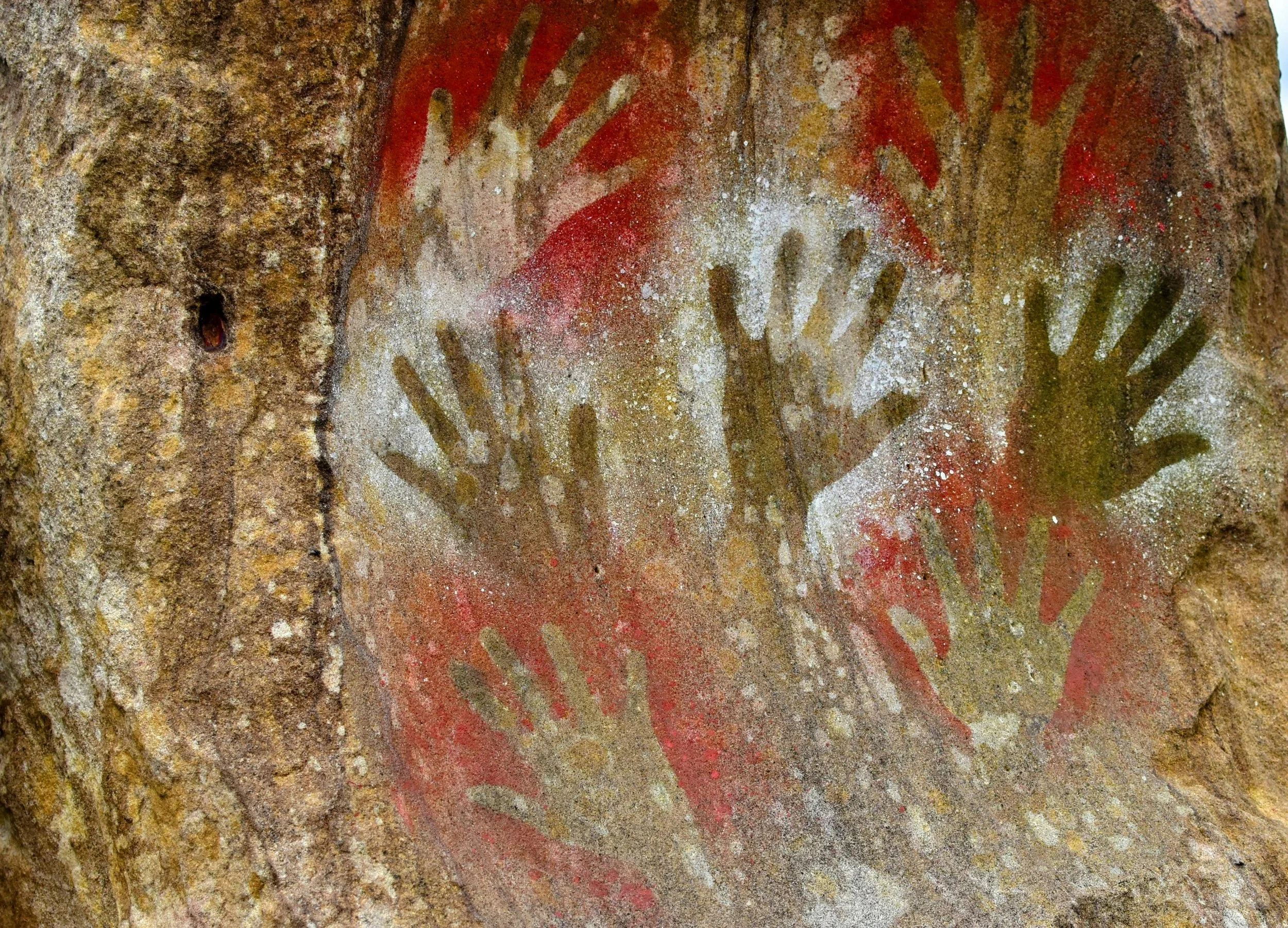 Ancient cave painting of multiple handprints in red and white on a rock surface.