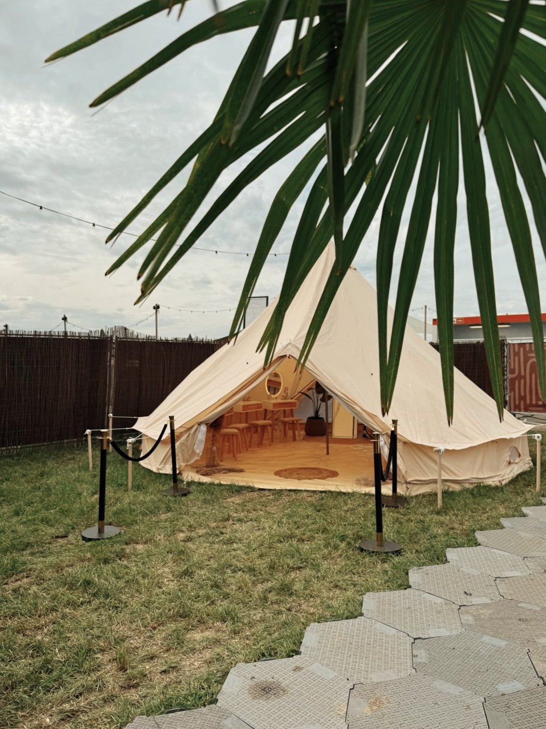 Ein Zelt im Freien, sichtbar durch eine große grüne Palmenblatt im Vordergrund, mit Stühlen und einem Teppich im Inneren, umgeben von einer schwarzen Absperrung und einer Gartenumgebung.