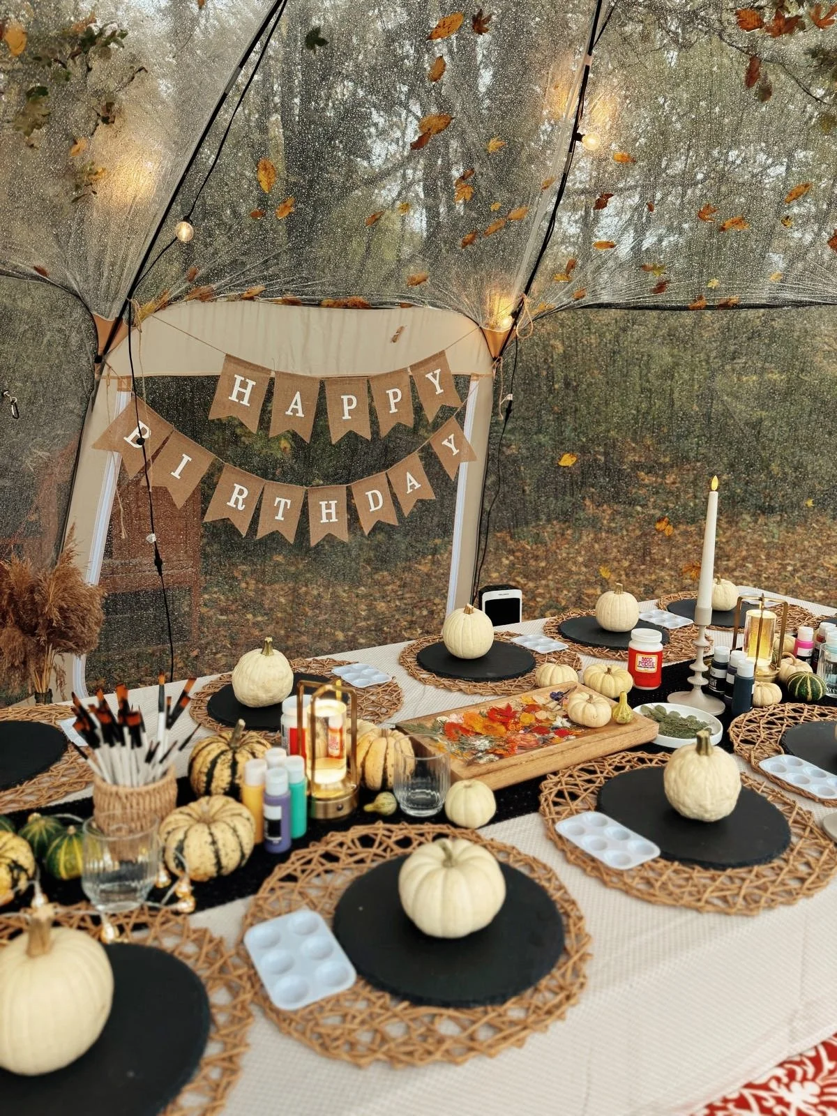 Tischdekoration mit weißen Kürbissen, Herbstblättern, Kerzen und Halloween-Farben, für eine Geburtstagsfeier, im Zelt mit einem 'Happy Birthday'-Banner im Hintergrund.