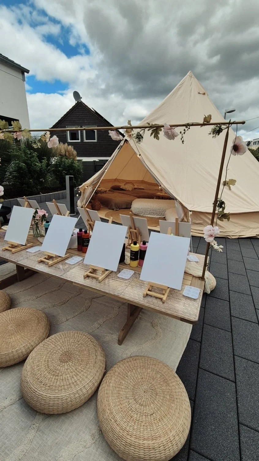 Ein Outdoor-Achtzelt mit Bett und Kissen, umgeben von einer dekorativen Blumenleiste, auf einem Dachgarten. Vor dem Zelt steht ein Tisch mit Malutensilien und Leinwänden, umgeben von Sitzkissen.