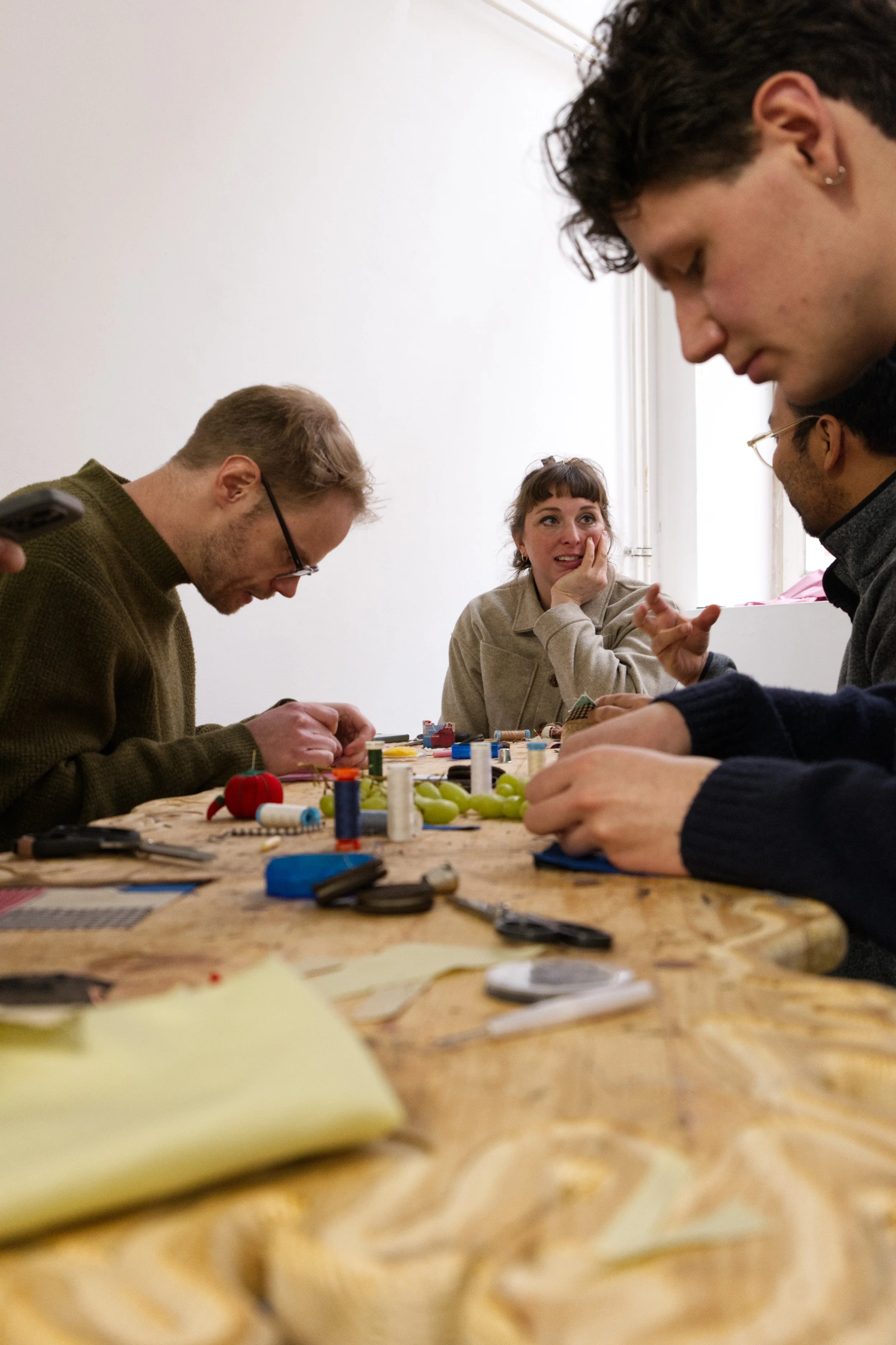 3 man and a woman seated at a wooden table, doing needle work and being in conversation.