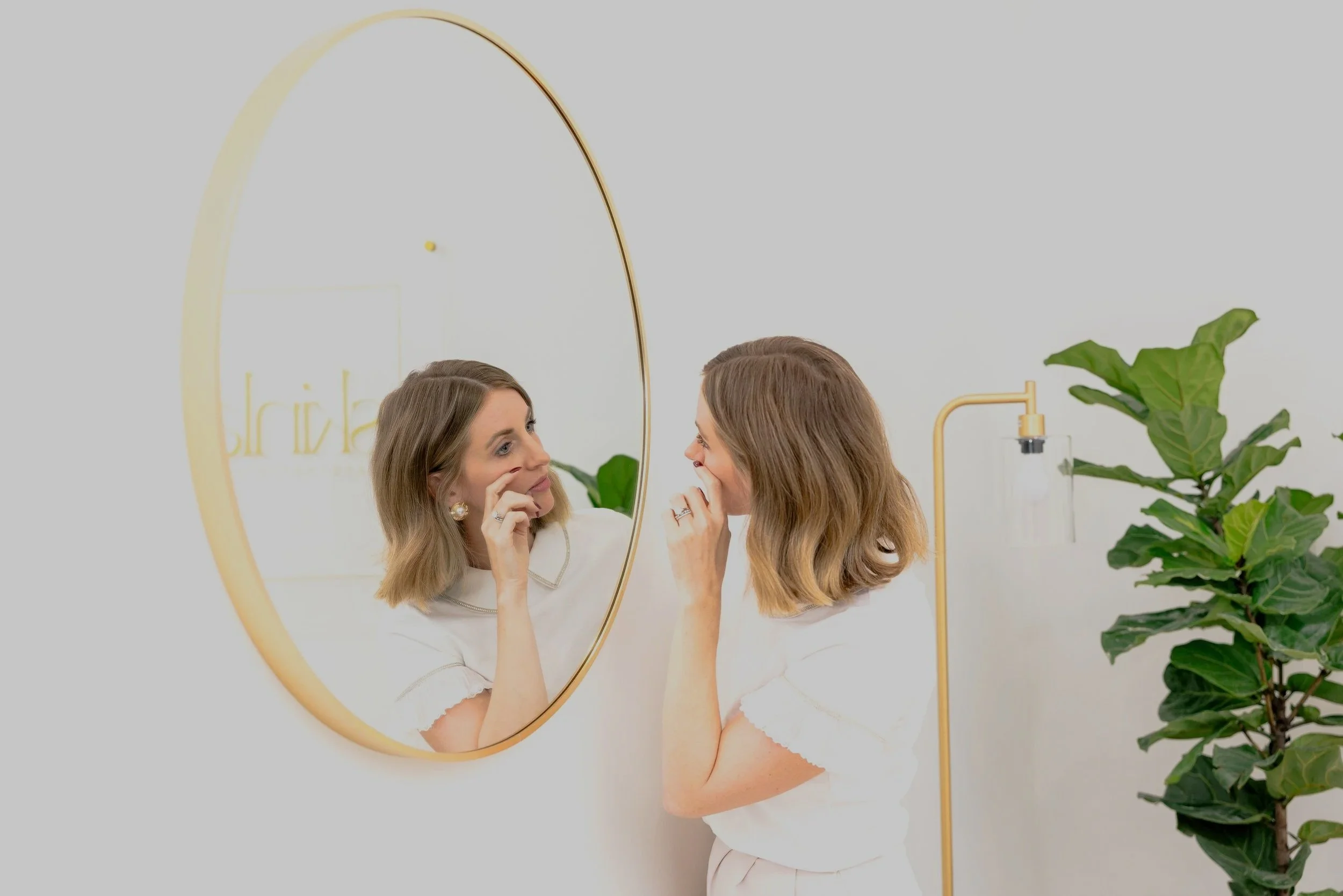 A woman with blonde hair looking at her reflection in a round mirror, touching her face, in a bright, modern room with green plants and minimal decor.