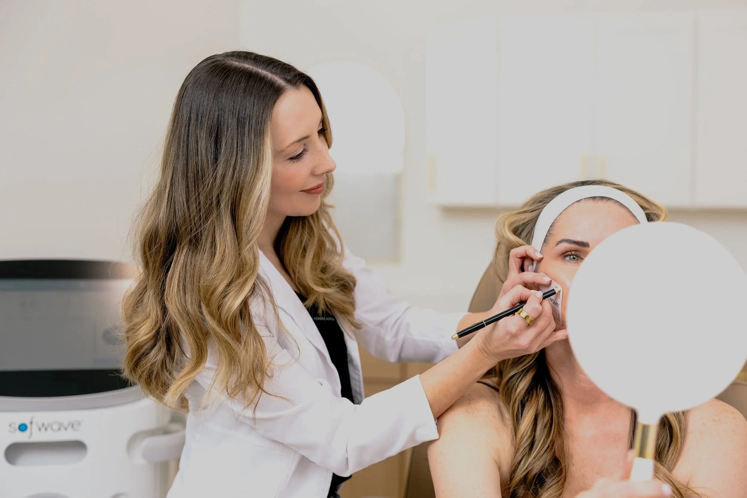 A woman with long, wavy blonde hair is receiving a cosmetic or dermatological treatment from a professional in a clinical setting. The professional has long, light brown hair with blonde highlights and is wearing a white coat. The patient is holding a mirror while the professional appears to be marking or drawing on her face.