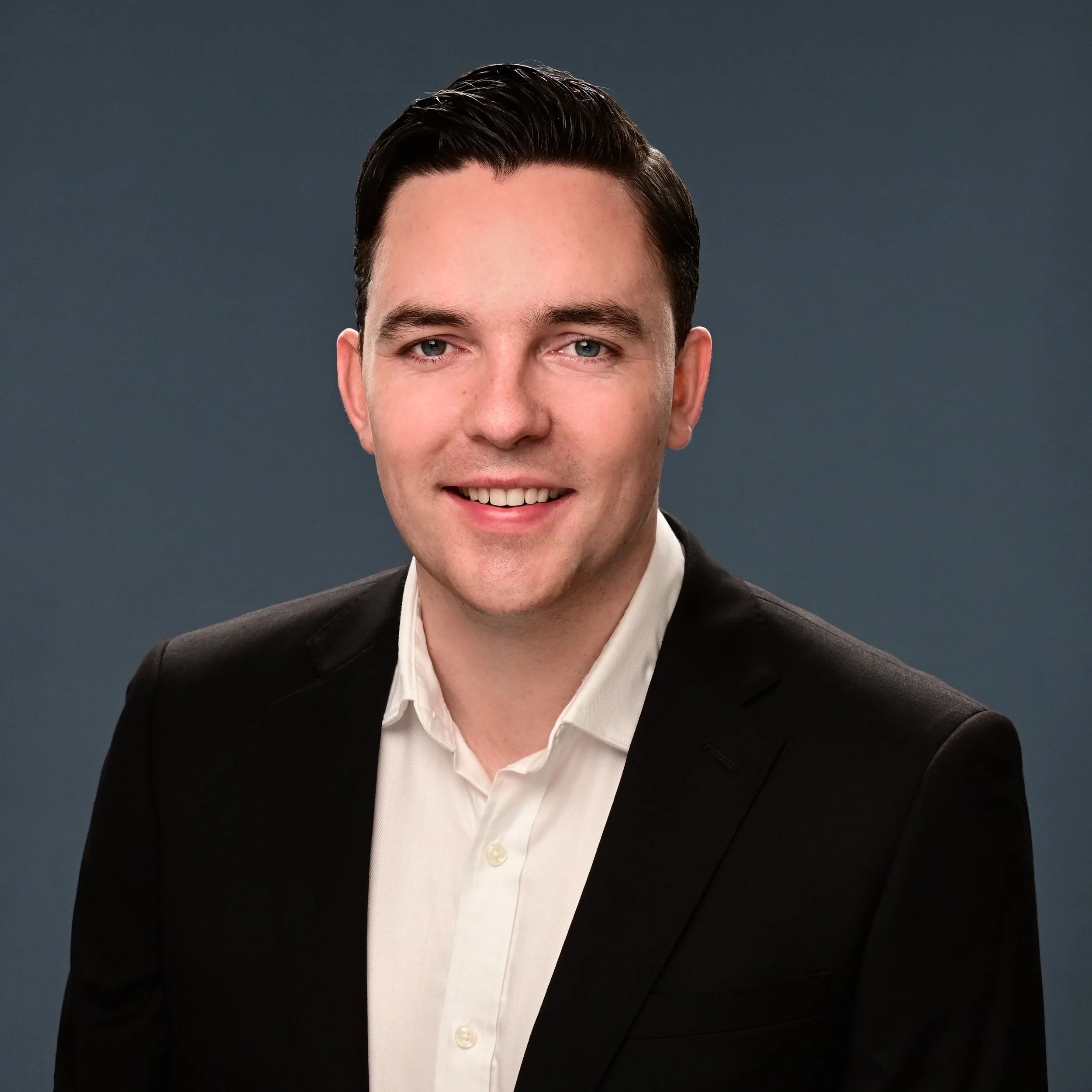Professional headshot of a smiling man dressed in a black suit and white shirt against a dark gray background.