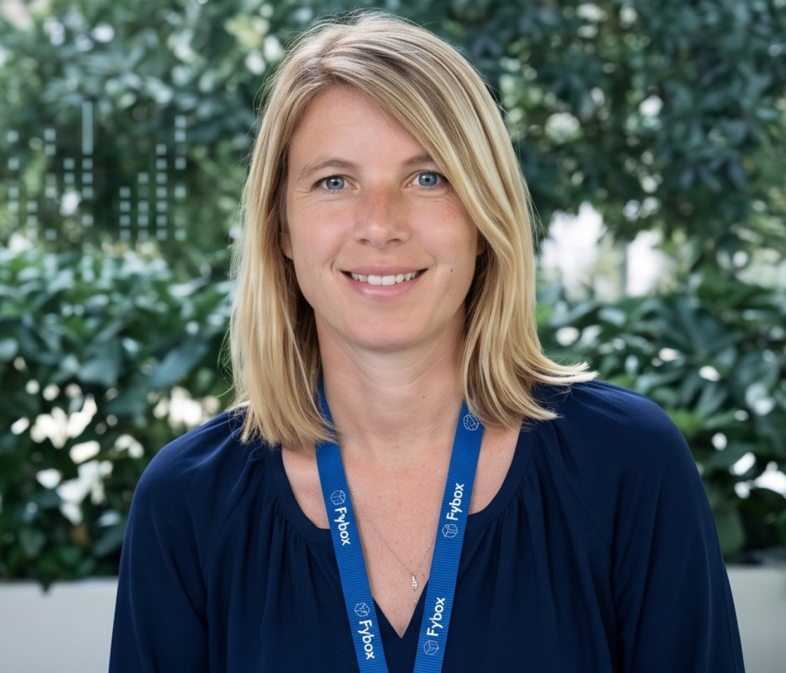 Portrait of a woman with blonde hair, blue eyes, wearing a navy blue top and a blue lanyard with 'Flybox' written on it, standing in front of green foliage.