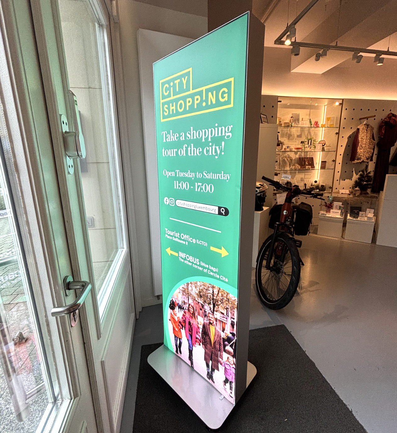 Digital signboard promoting city shopping tours in Luxembourg, displaying opening hours, social media icons, and directions to tourist office and info bus on a turquoise background with an image of tourists at the bottom.