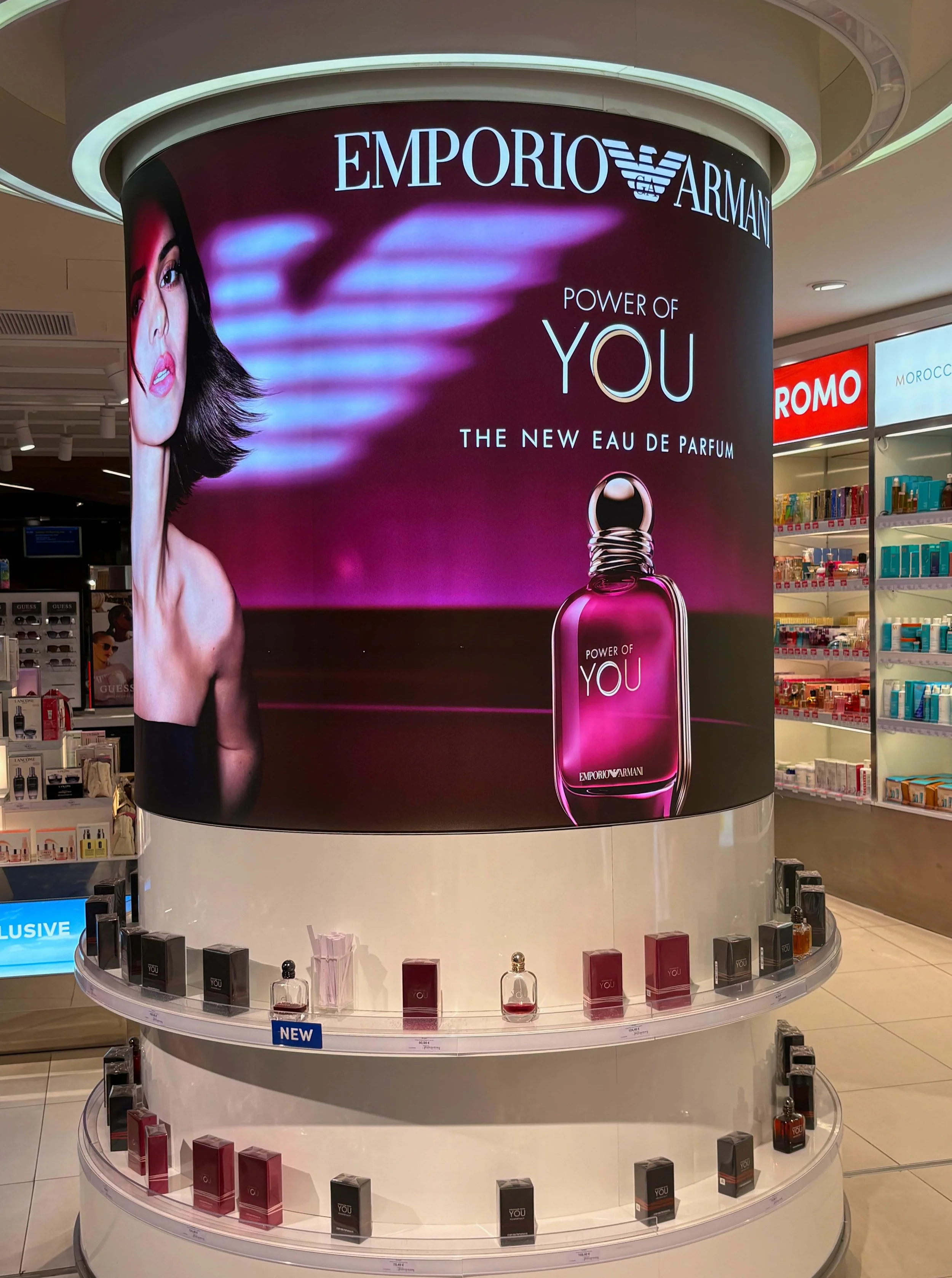 A large circular advertisement for Emporio Armani's fragrance 'Power of You' in a shopping mall. The ad features a woman with dark hair and bold makeup on the left side, and a pink perfume bottle on the right side. Below the ad, there are shelves displaying various perfume bottles and boxes.