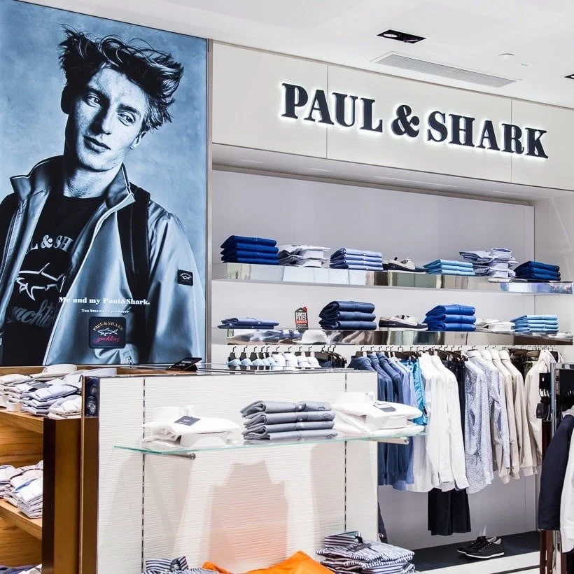 Interior of a clothing store named Paul & Shark with folded shirts and hanging garments, a large black and white poster of a woman, and the store's sign.