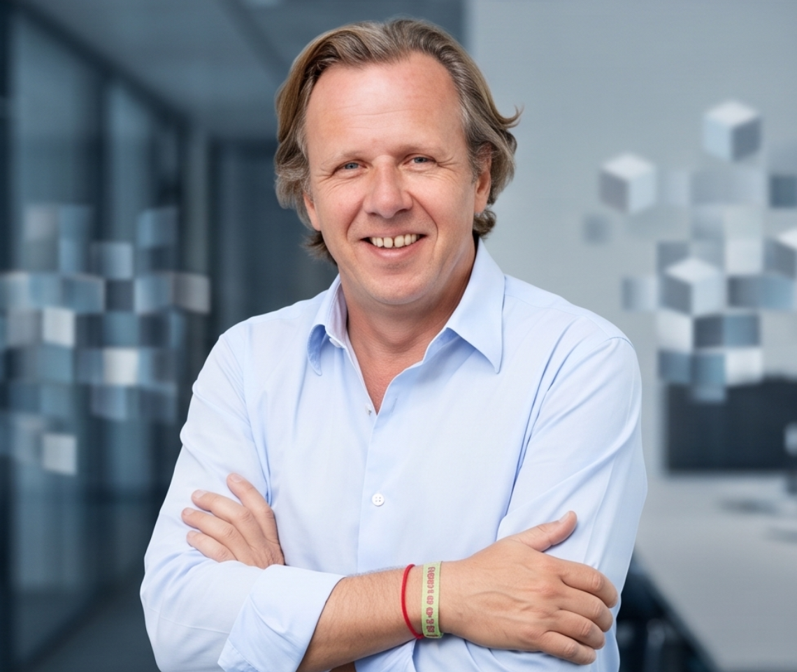 A smiling middle-aged man with blonde hair, wearing a light blue shirt, standing in a modern office with abstract decor in the background.
