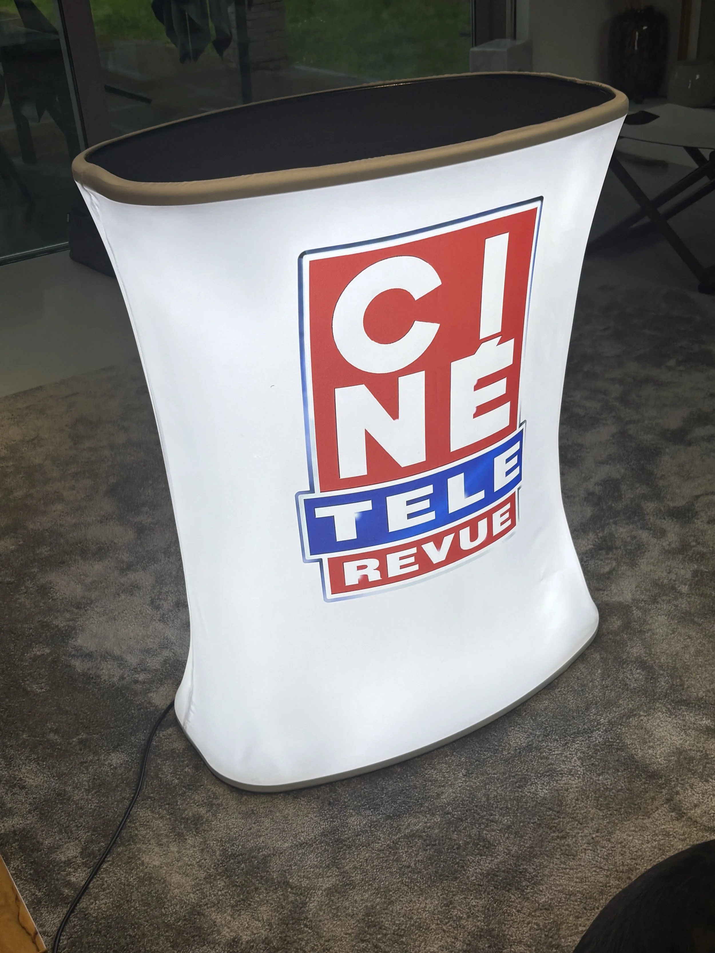 A portable sign with the logos of a convenience store called 'CONE', with the words 'TULE REVU' underneath, illuminated from within, standing on a carpeted floor.