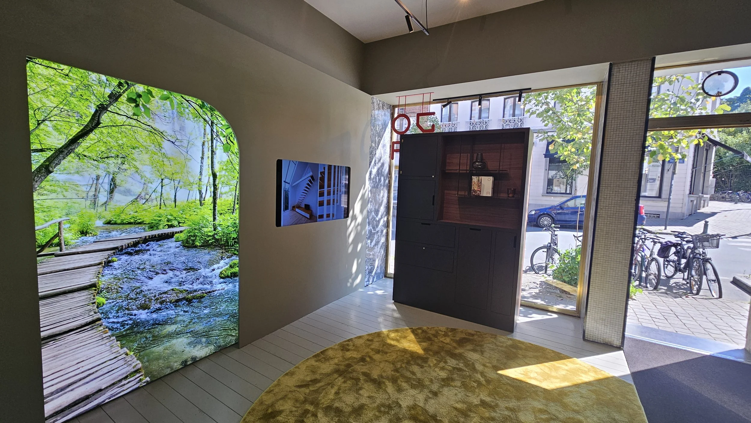 Interior of a modern space with a large nature-themed mural of a forest stream on the left wall, a faucet-shaped item on a dark wooden shelf, large windows showing bicycles outside, and a yellow circular rug on white wooden flooring.