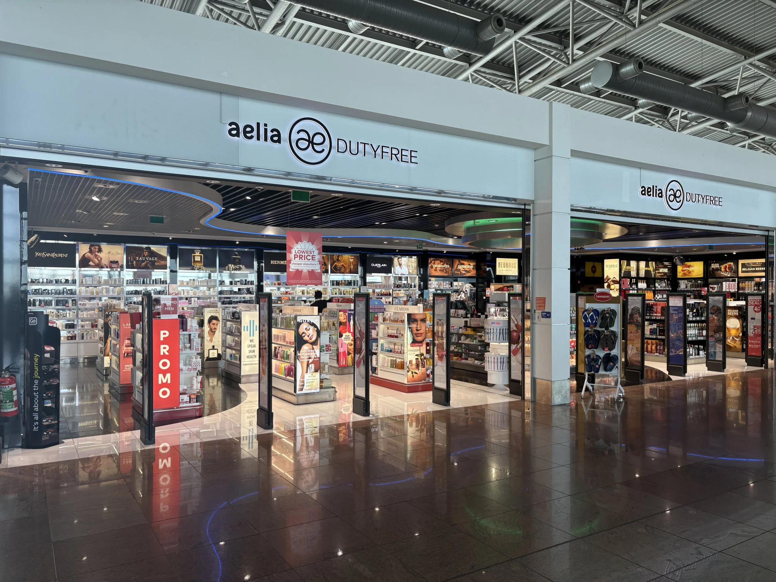 Interior of an airport duty-free shop with various products displayed on shelves, including perfumes, cosmetics, and accessories, with a lit sign that reads 'aelia duty free' above the entrance.