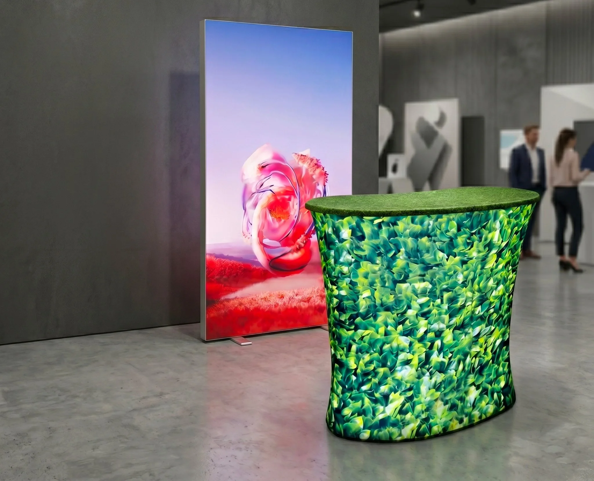 An exhibition display with a vertical digital screen showing colorful abstract art, and a rounded table with a green leafy pattern in the foreground.