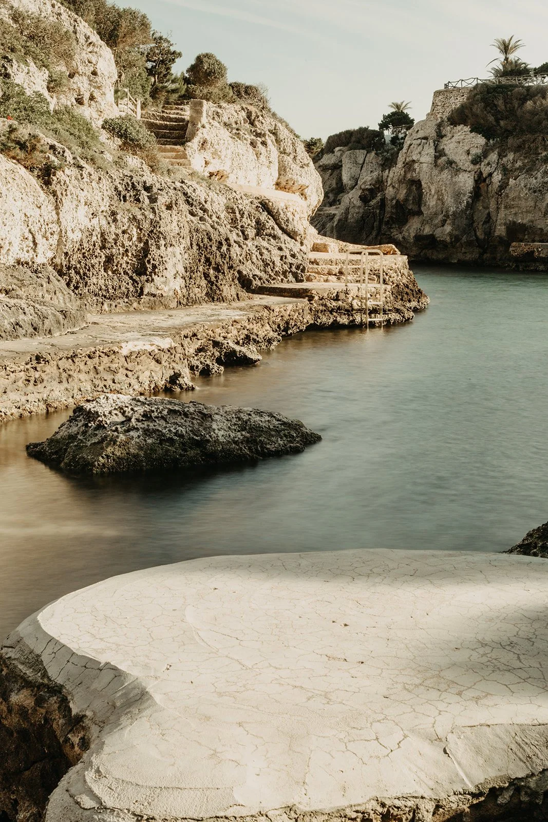 Quiet cala in Menorca with stairs leading into water, surrounded by sparse vegetation, under a clear sky.