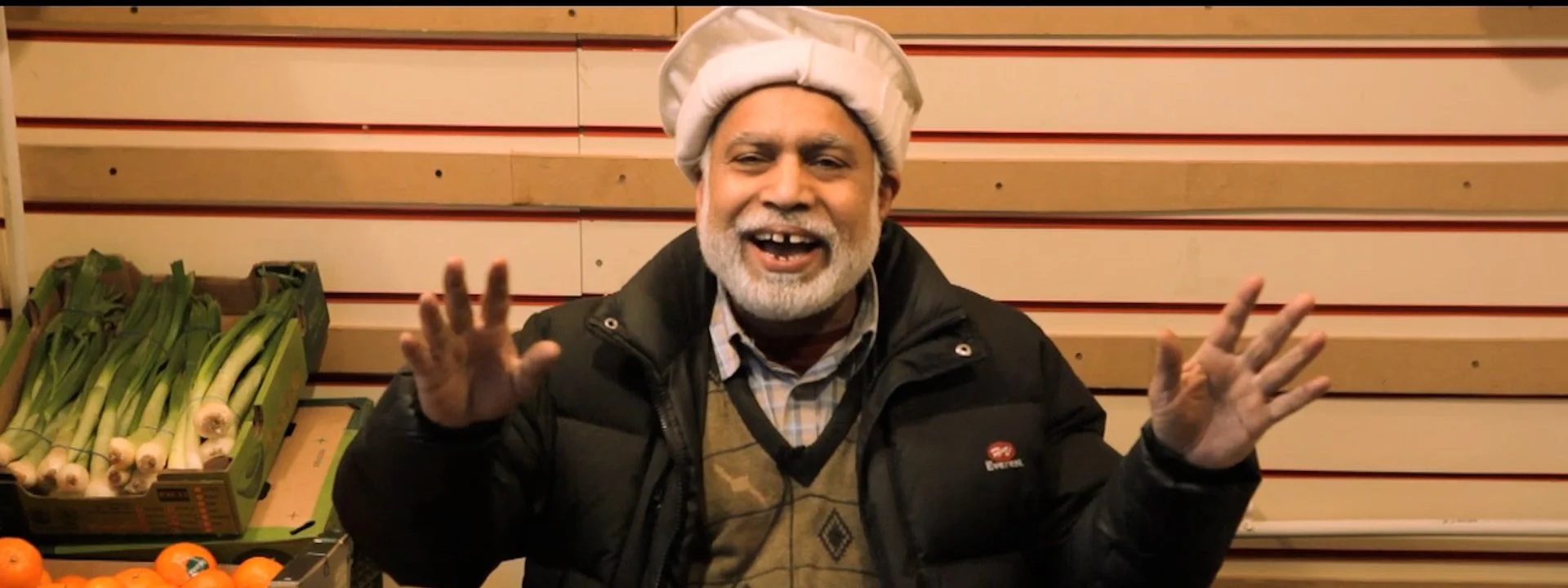 A man with gray beard wearing a white cap, black jacket, and plaid shirt, smiling and waving at the camera inside a store with various vegetables in the background.
