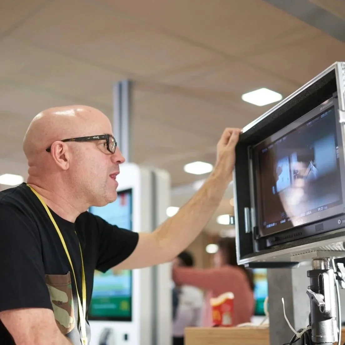 A bald man with glasses looking at a monitor in a busy indoor setting.