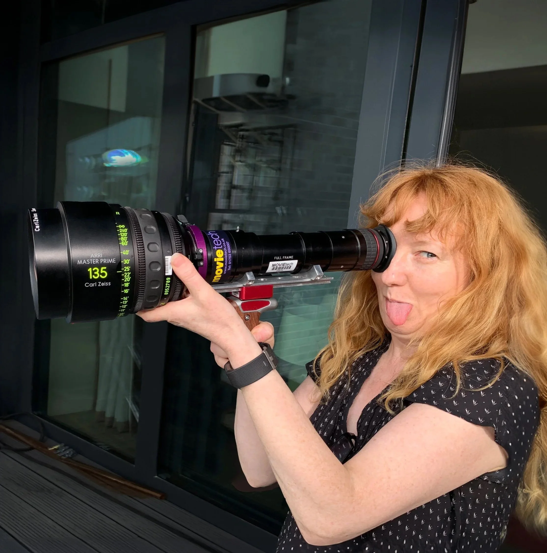 Film director Ita FitzGerald, with curly red hair, looks through a large camera lens viewfinder while sticking out her tongue.