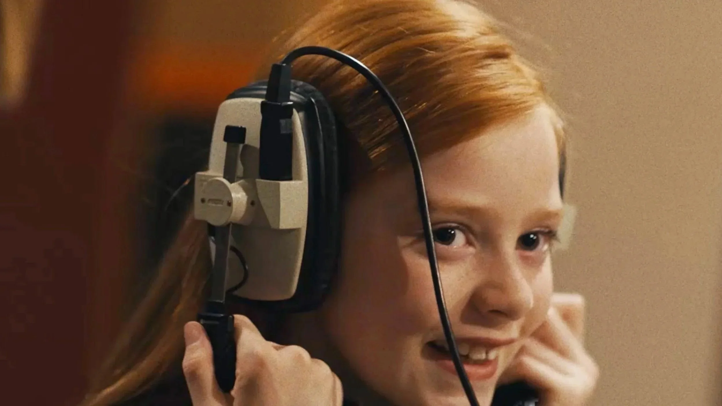 A young girl with red hair listening with headphones, smiling and holding the headphone cord.