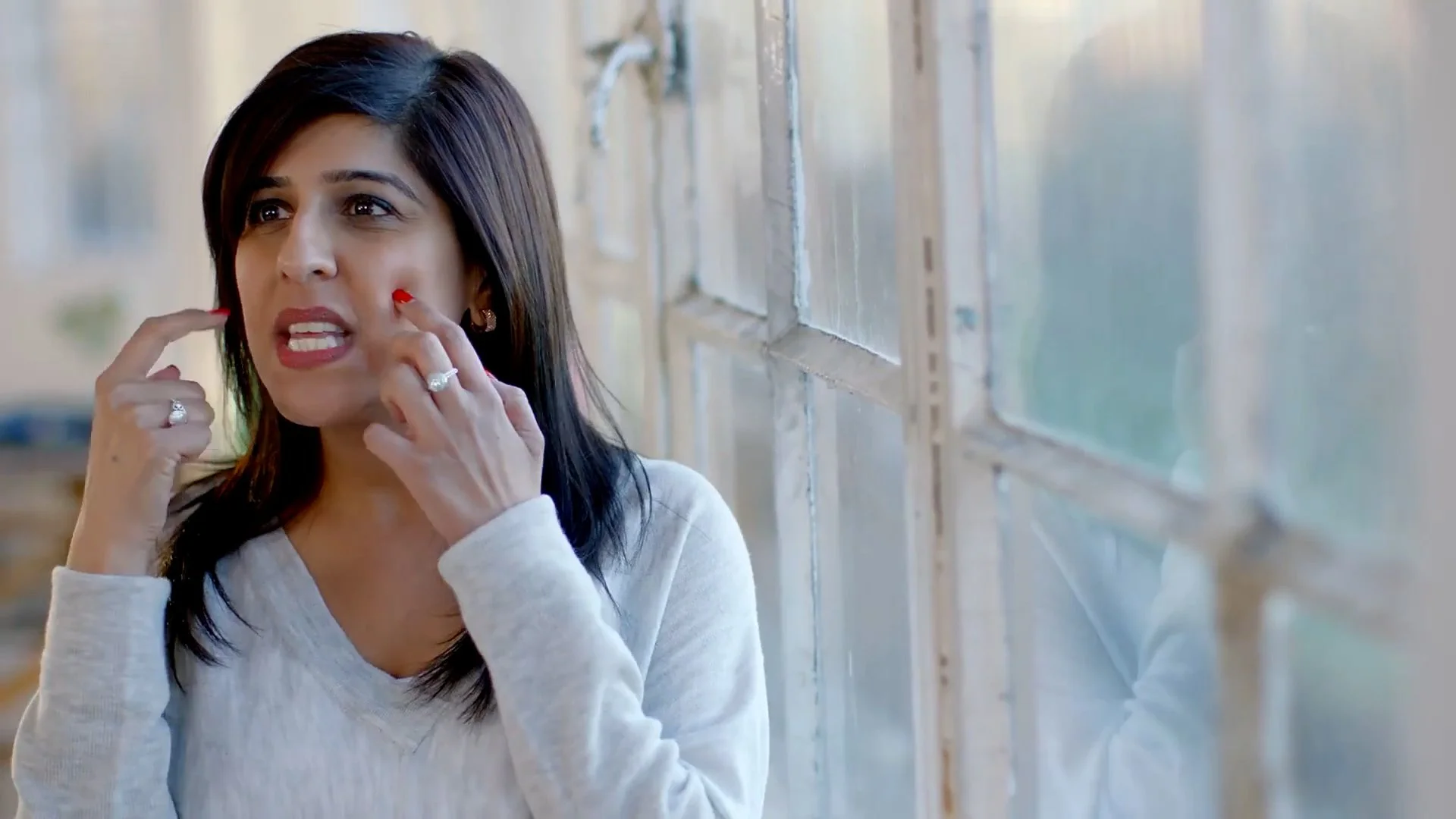 A woman with dark hair and wearing a light gray long-sleeve shirt is touching her face and appears to be anxious or upset while standing near a window.