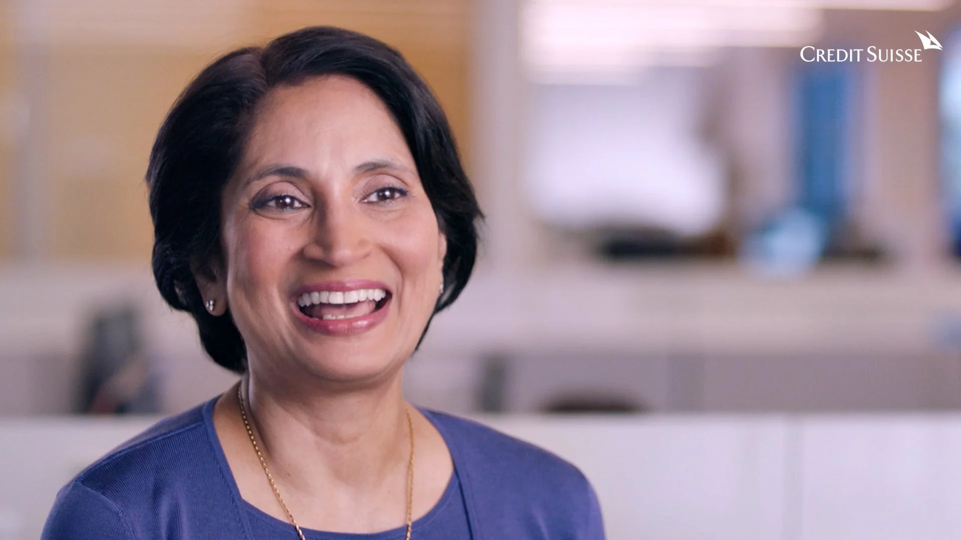 A smiling woman with short black hair, wearing earrings and a blue top, in an indoor setting with books in the background, with Credit Suisse logo in the top right corner.