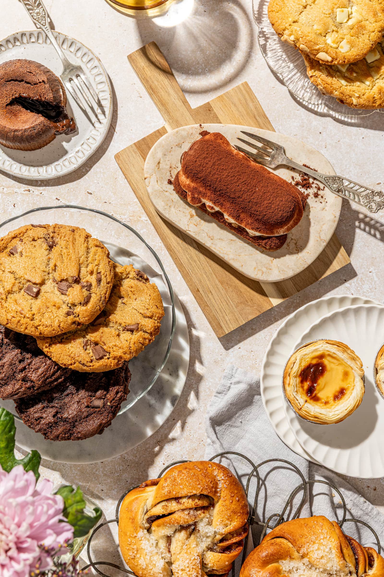 Assorted cookies, pastries, and desserts on plates and a wooden board, with a pink flower arrangement partially visible in the corner.