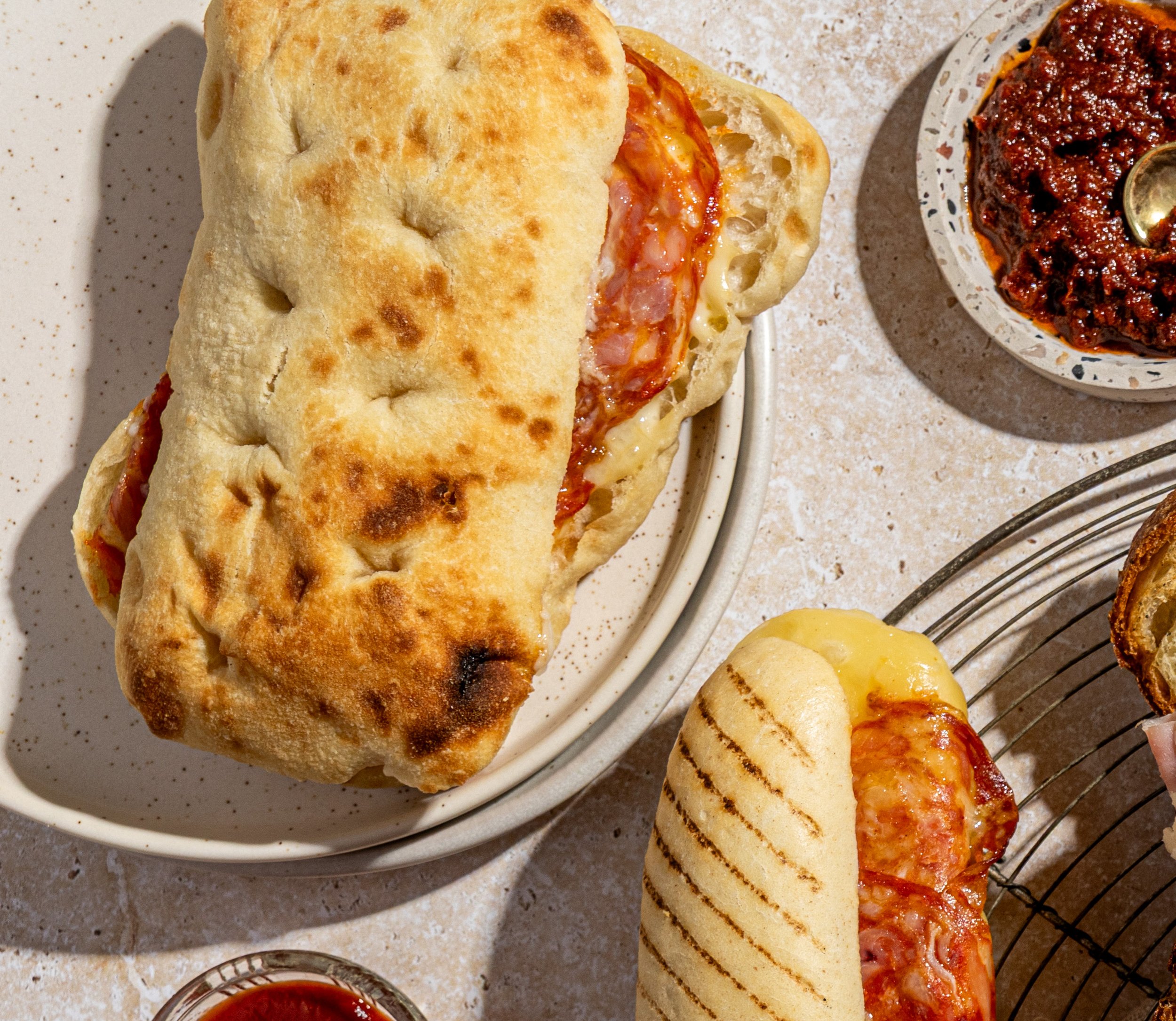 Two paninis on plates, melted cheese and salami, accompanied by small bowls of sauce or tomato paste on a speckled countertop.