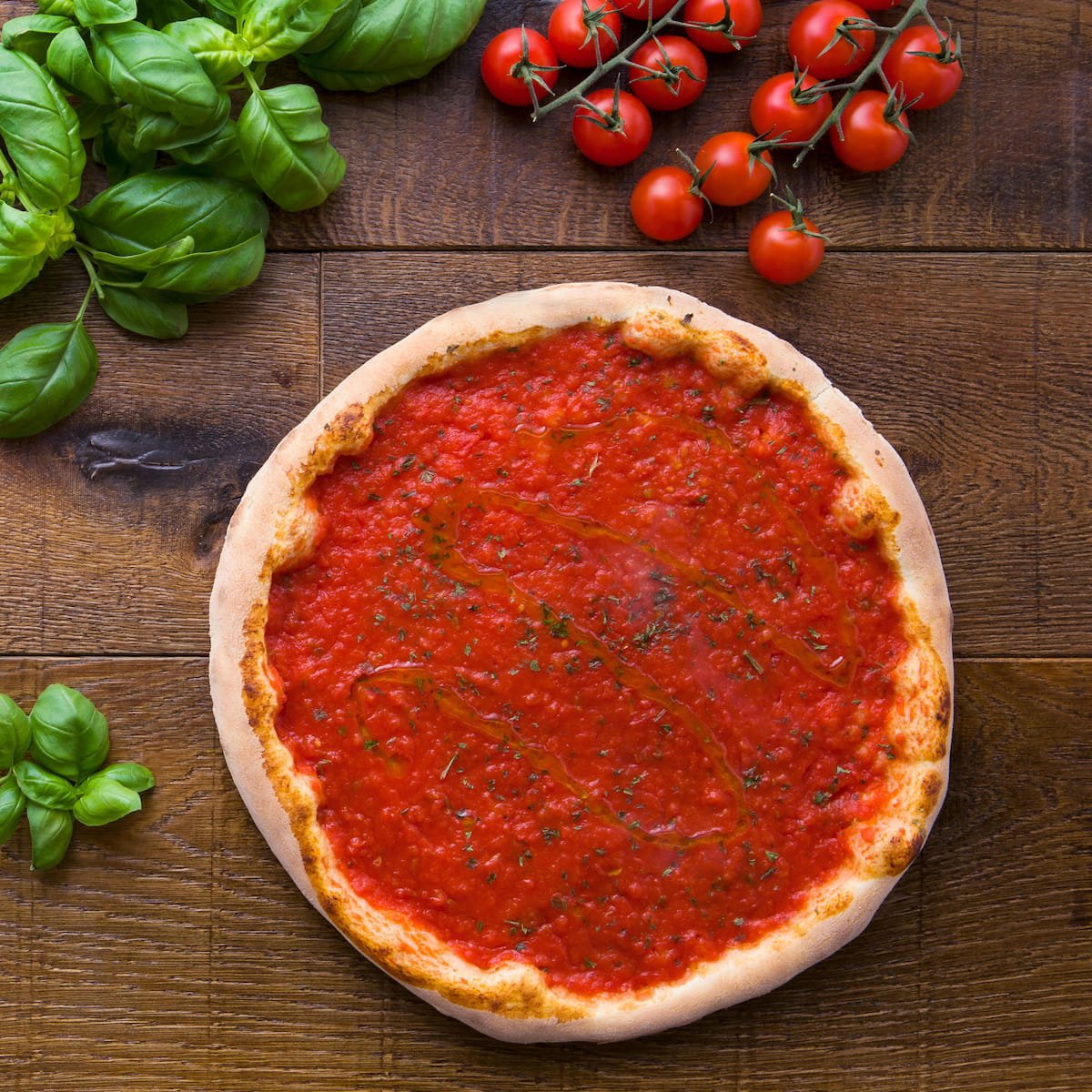 A pizza with tomato sauce, placed on a wooden surface with fresh basil leaves on the side and a bunch of cherry tomatoes nearby.