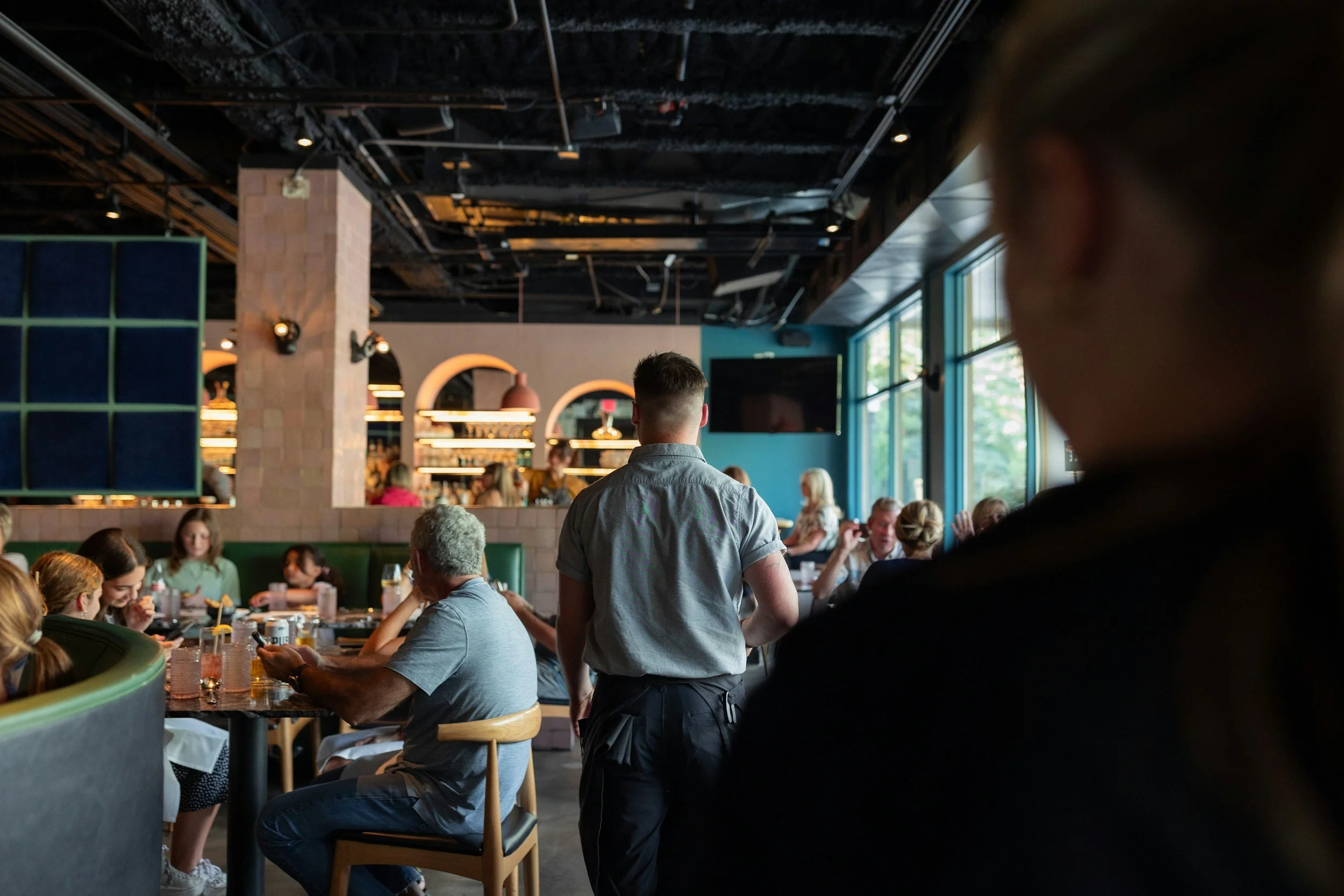 A group of people dining in a modern restaurant with natural light from large windows, a bartender working behind the bar, and a server walking through the dining area.