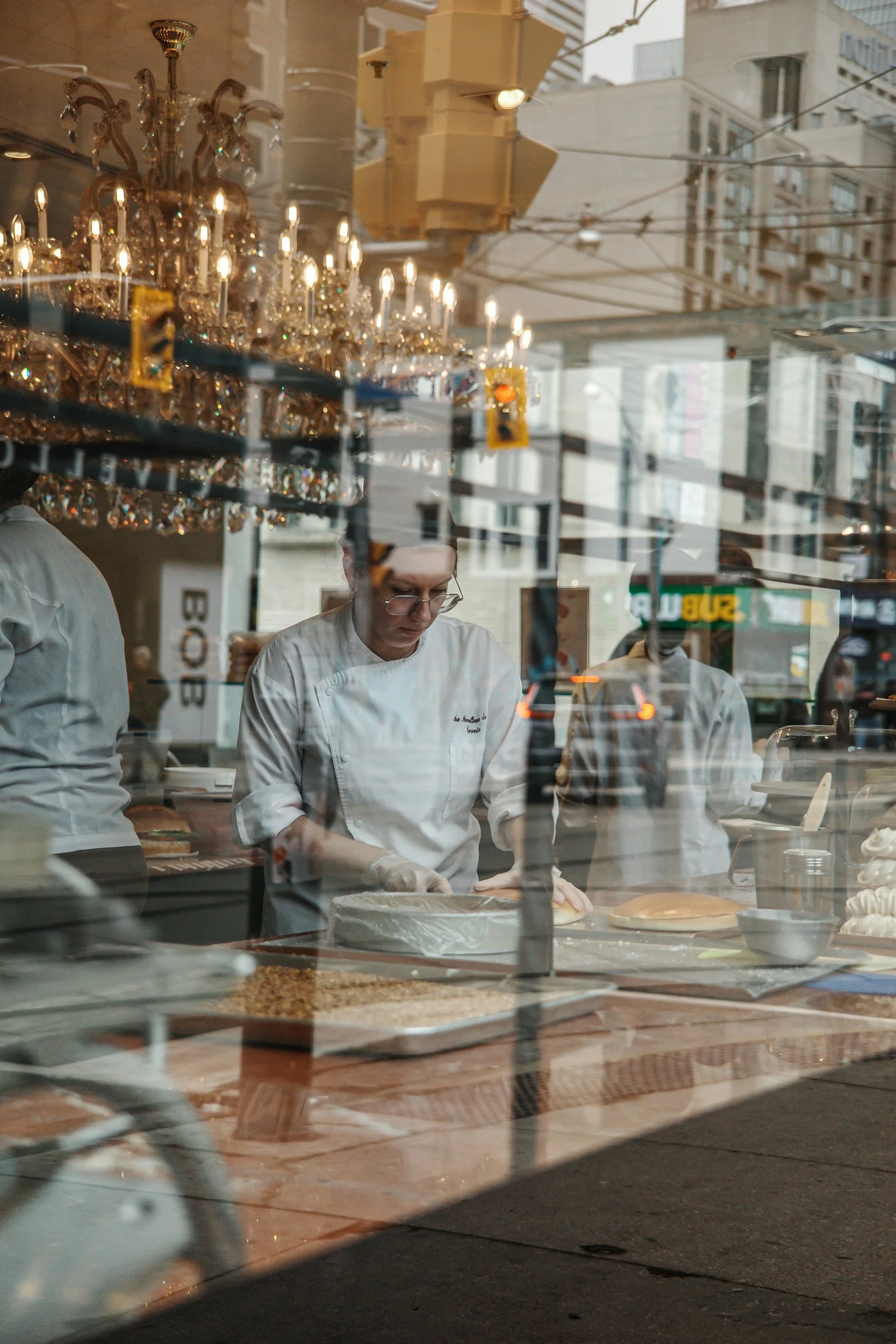 A chef appears to be preparing a dessert behind a shop window, with reflections of city buildings, traffic lights, and pedestrians outside.