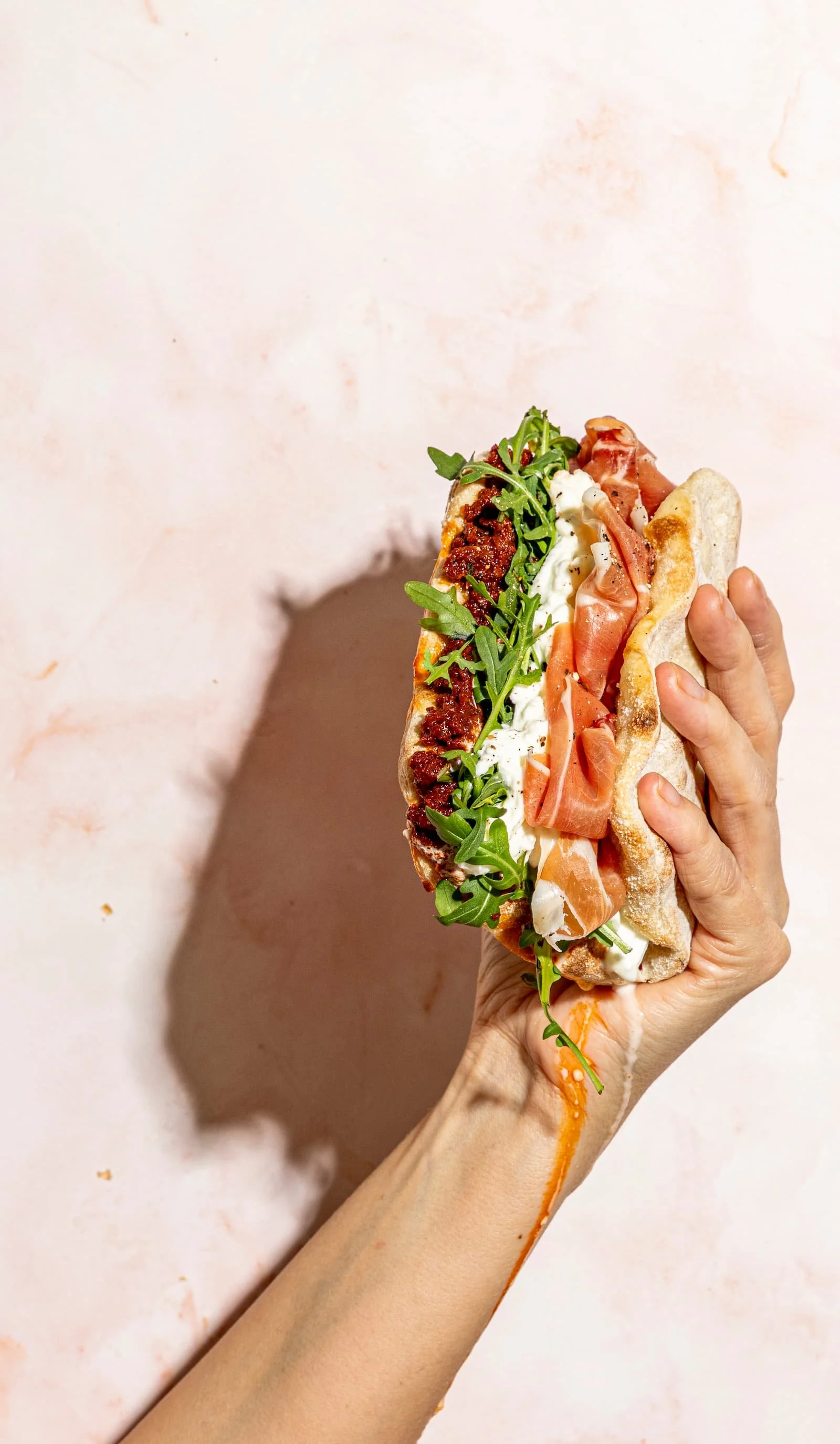 A hand holding a sandwich with arugula, smoked ham, mozzarella and pesto, against a light pink background.
